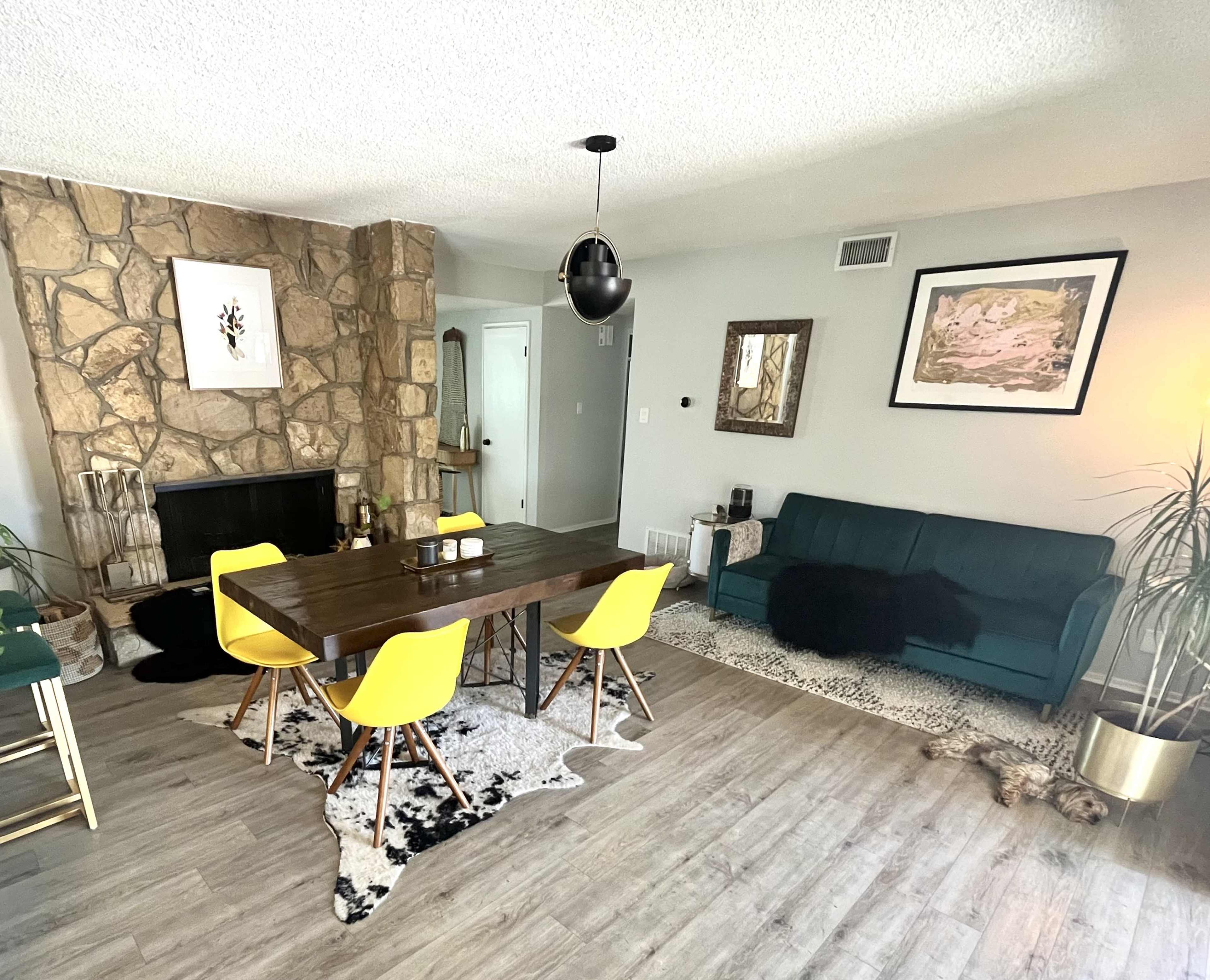 A modern living room with a stone fireplace, a wooden dining table surrounded by yellow chairs, and a green sofa against a light gray wall.