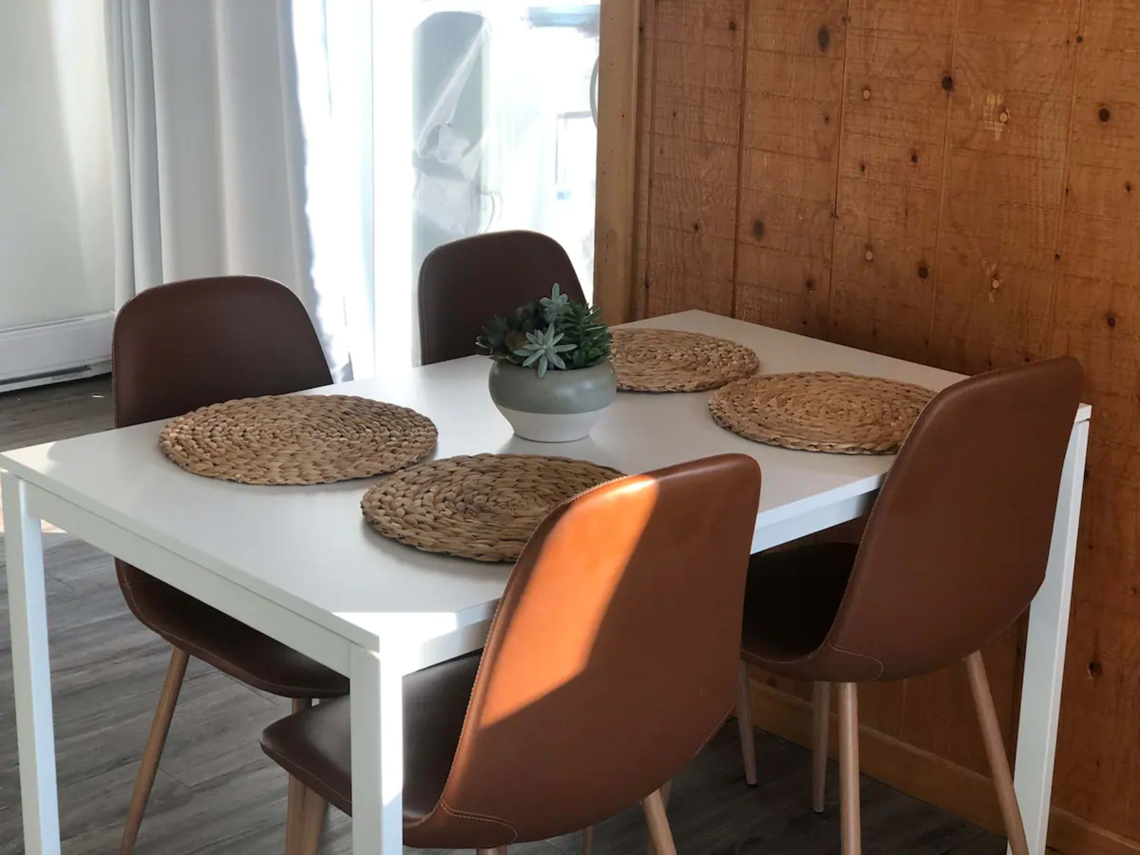 A white dining table is set with four brown chairs and round woven placemats, accompanied by a small potted plant at the center.