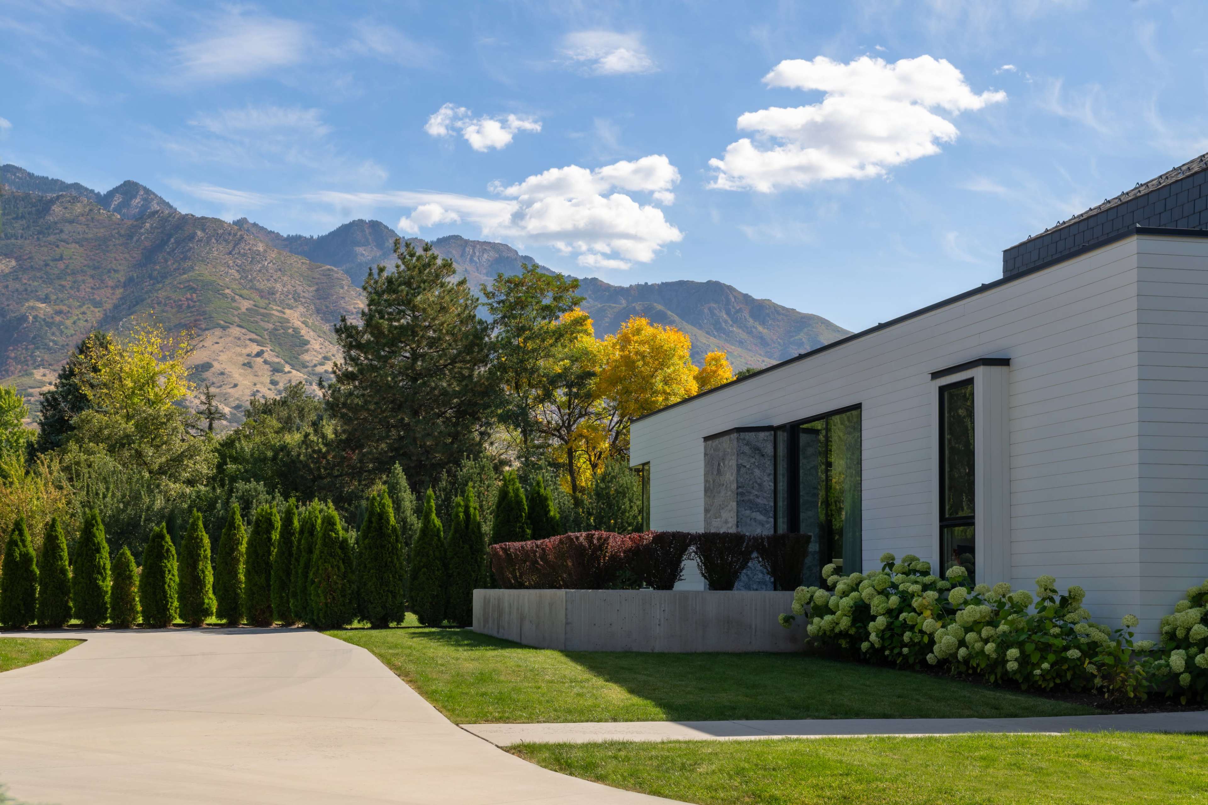 A modern house with large windows is situated on a driveway alongside neatly trimmed hedges and colorful trees, with mountains in the background.