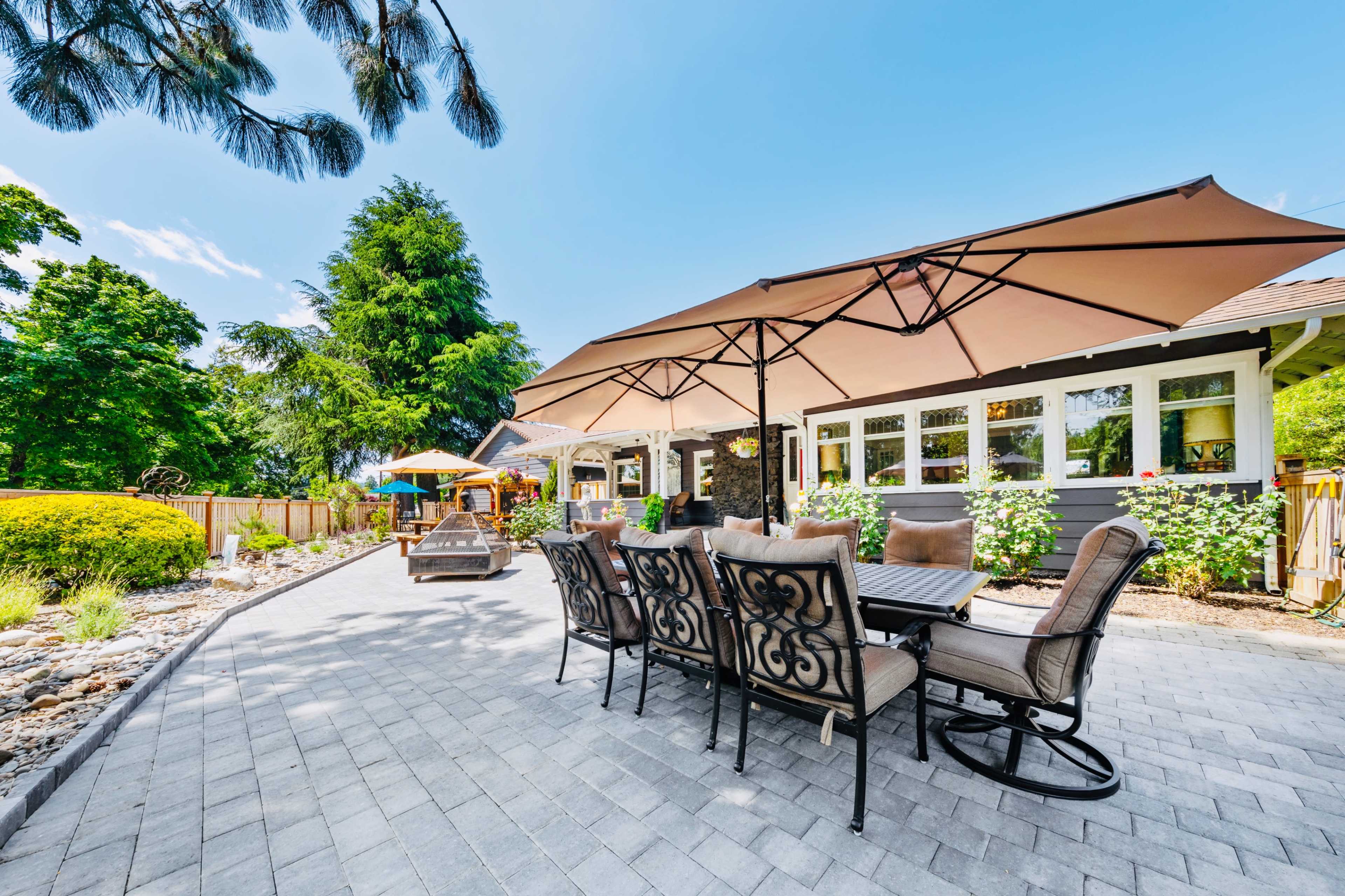 A patio area features a large dining set under beige umbrellas, surrounded by greenery and landscaped stones.