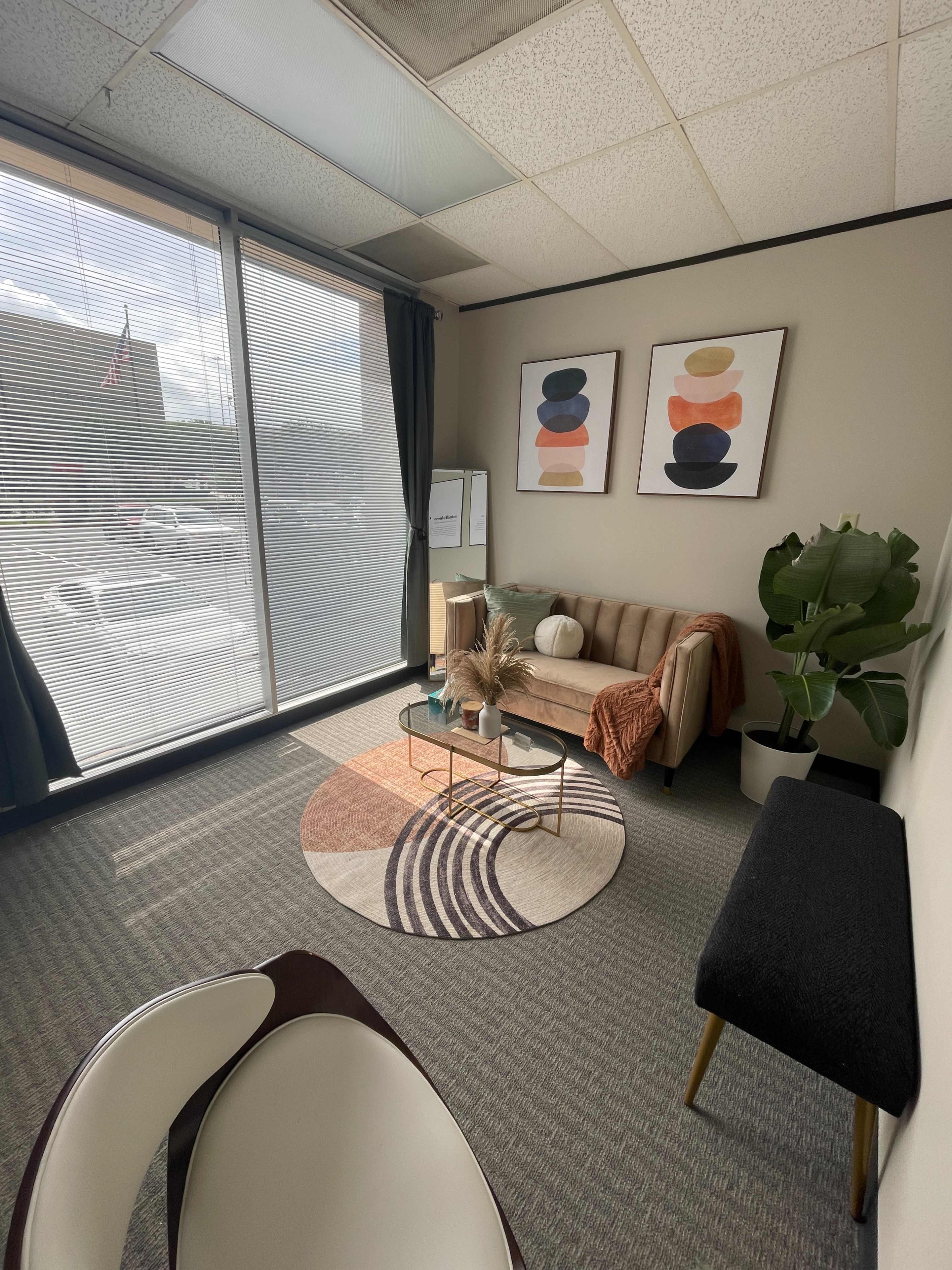 A well-lit office waiting area featuring a sofa, a coffee table, decorative plants, and two abstract art pieces on the wall, with large windows providing a view of the outside.