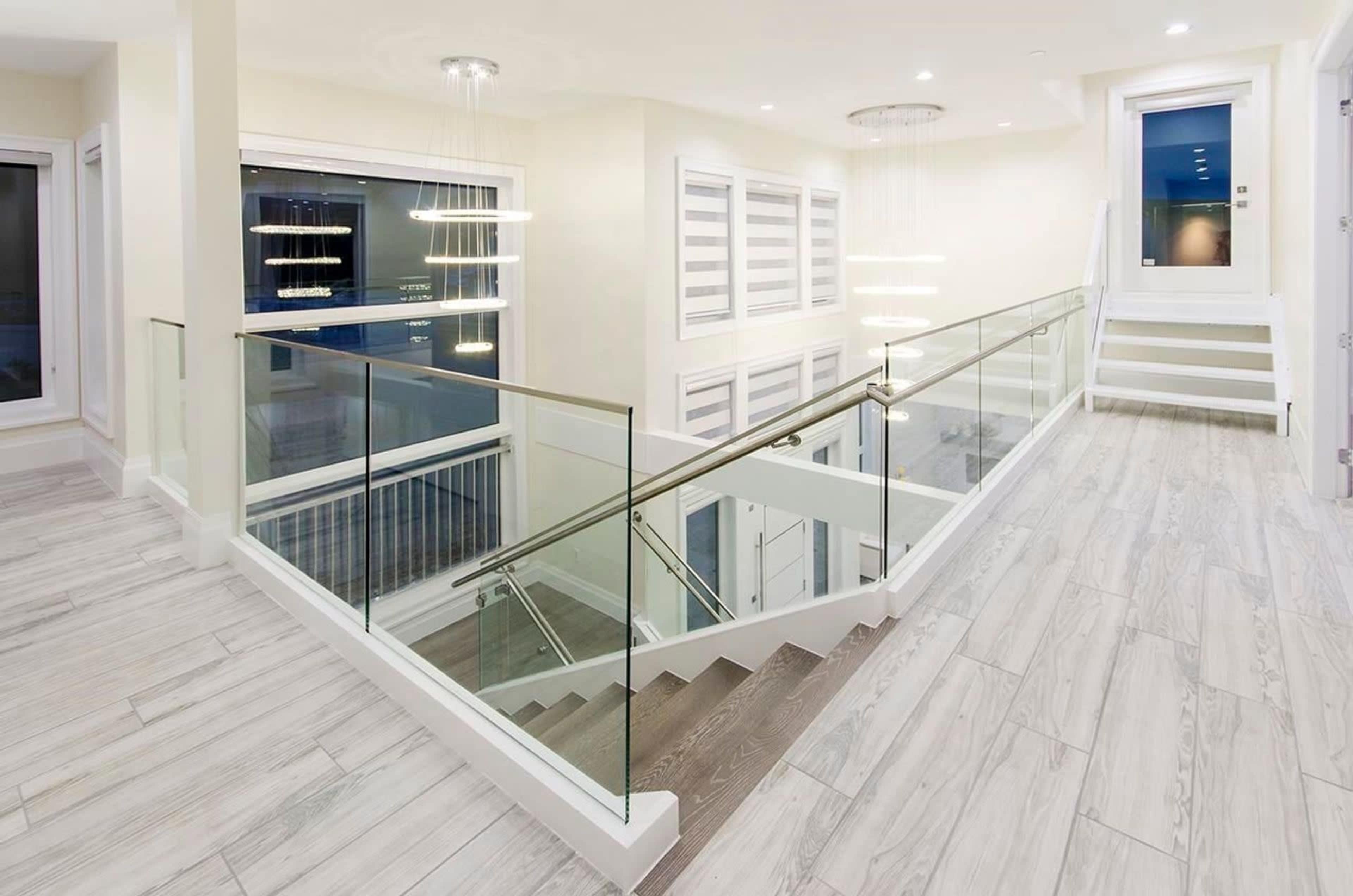A modern staircase with glass railings leading down to a well-lit living area, featuring large windows and contemporary lighting fixtures.