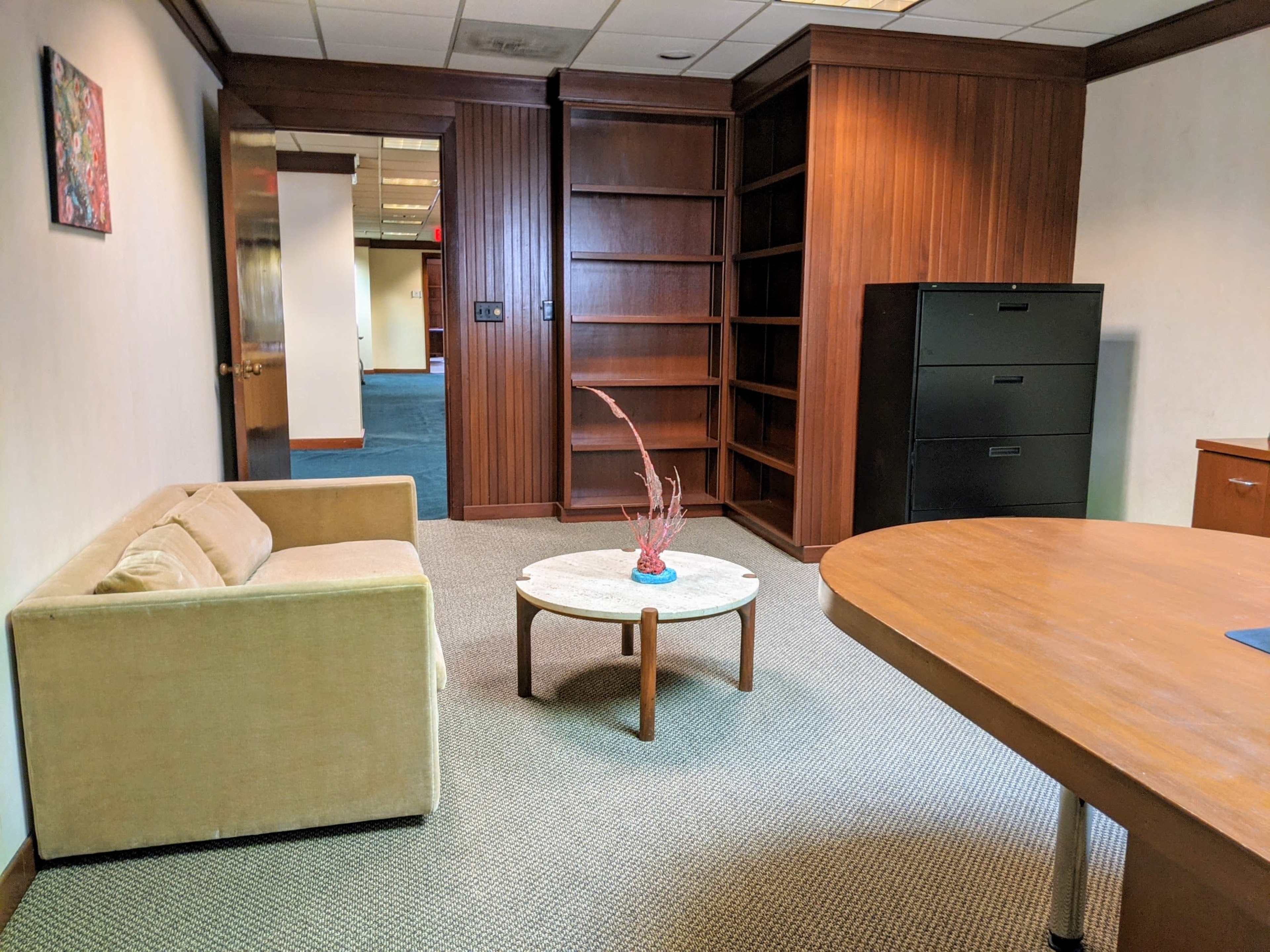 A small office space with a beige sofa, a round coffee table with a decorative plant, and a wooden meeting table, along with shelves and filing cabinets in the background.