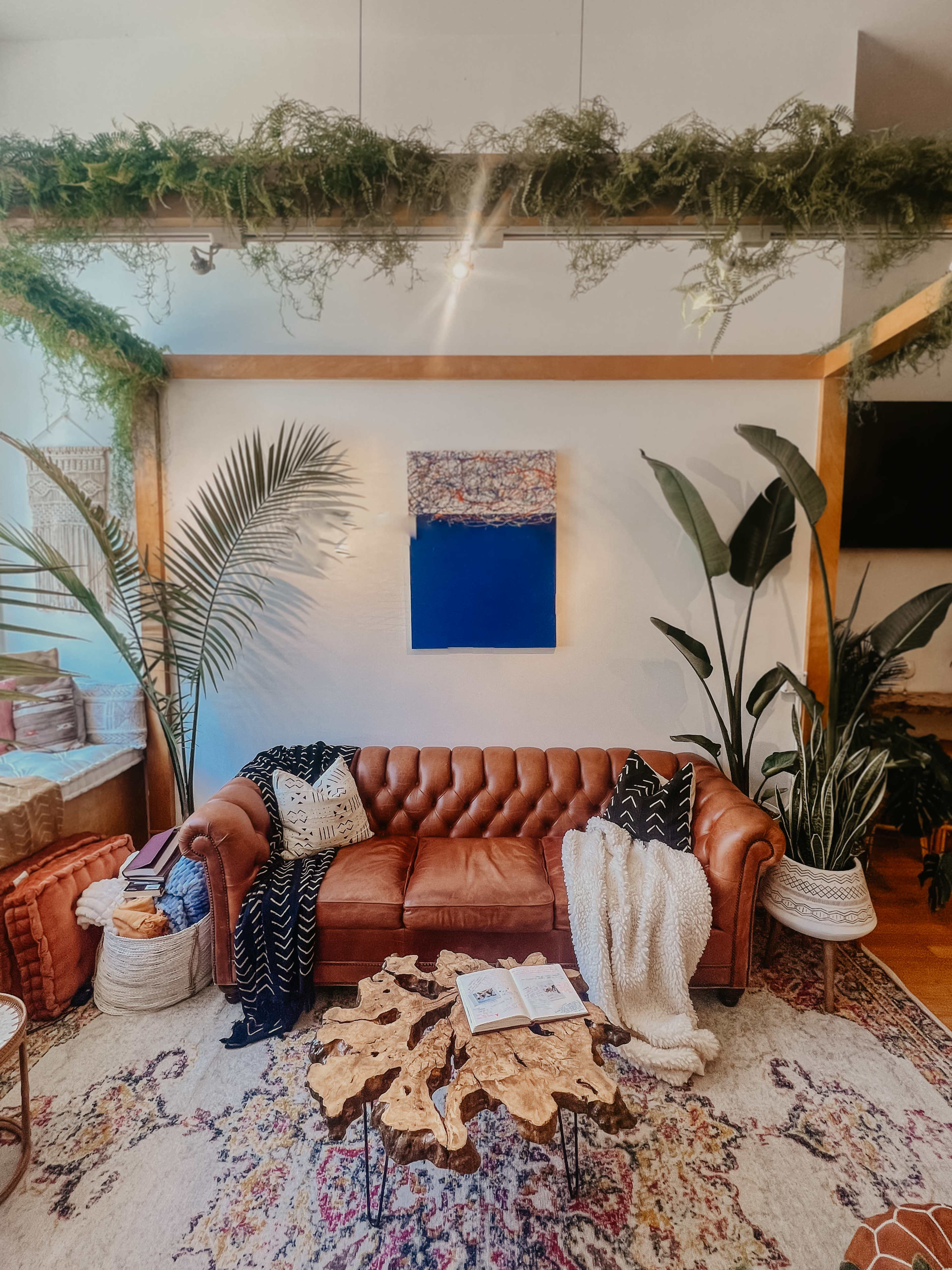 A brown leather couch is positioned in a bright living space decorated with plants, a colorful area rug, and a wooden coffee table with magazines on it.