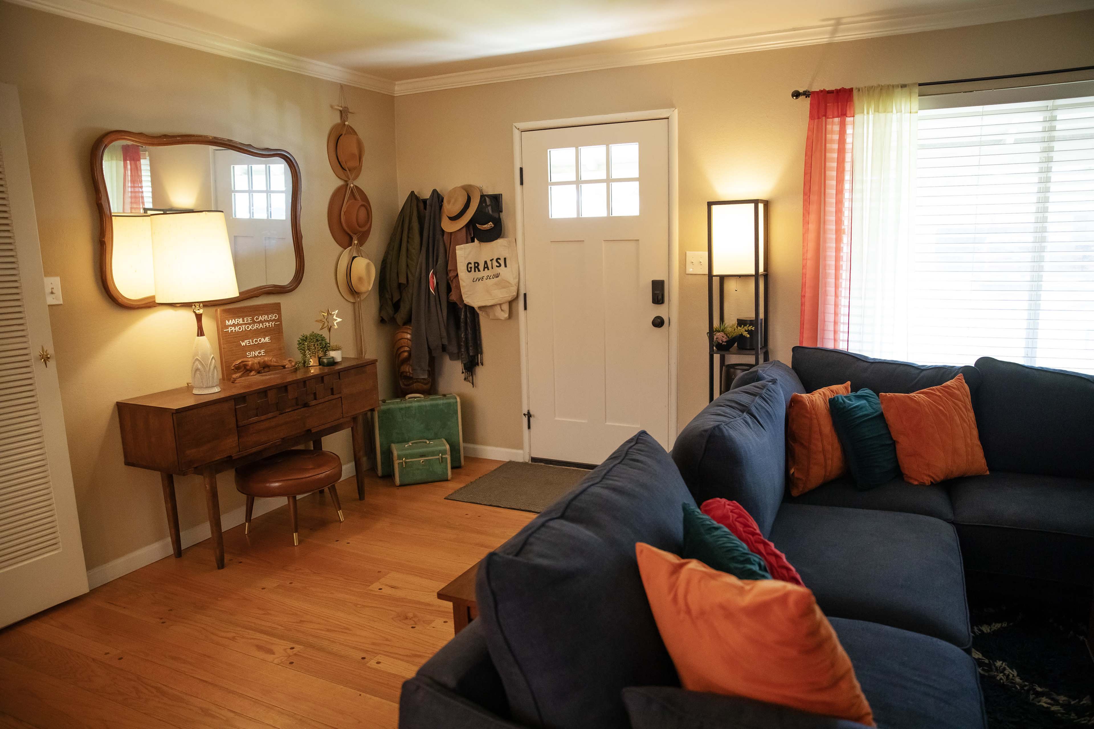A cozy living room features a blue sectional sofa with colorful cushions, a wooden entryway table with decorative items, and a door with window panes leading to the outside.