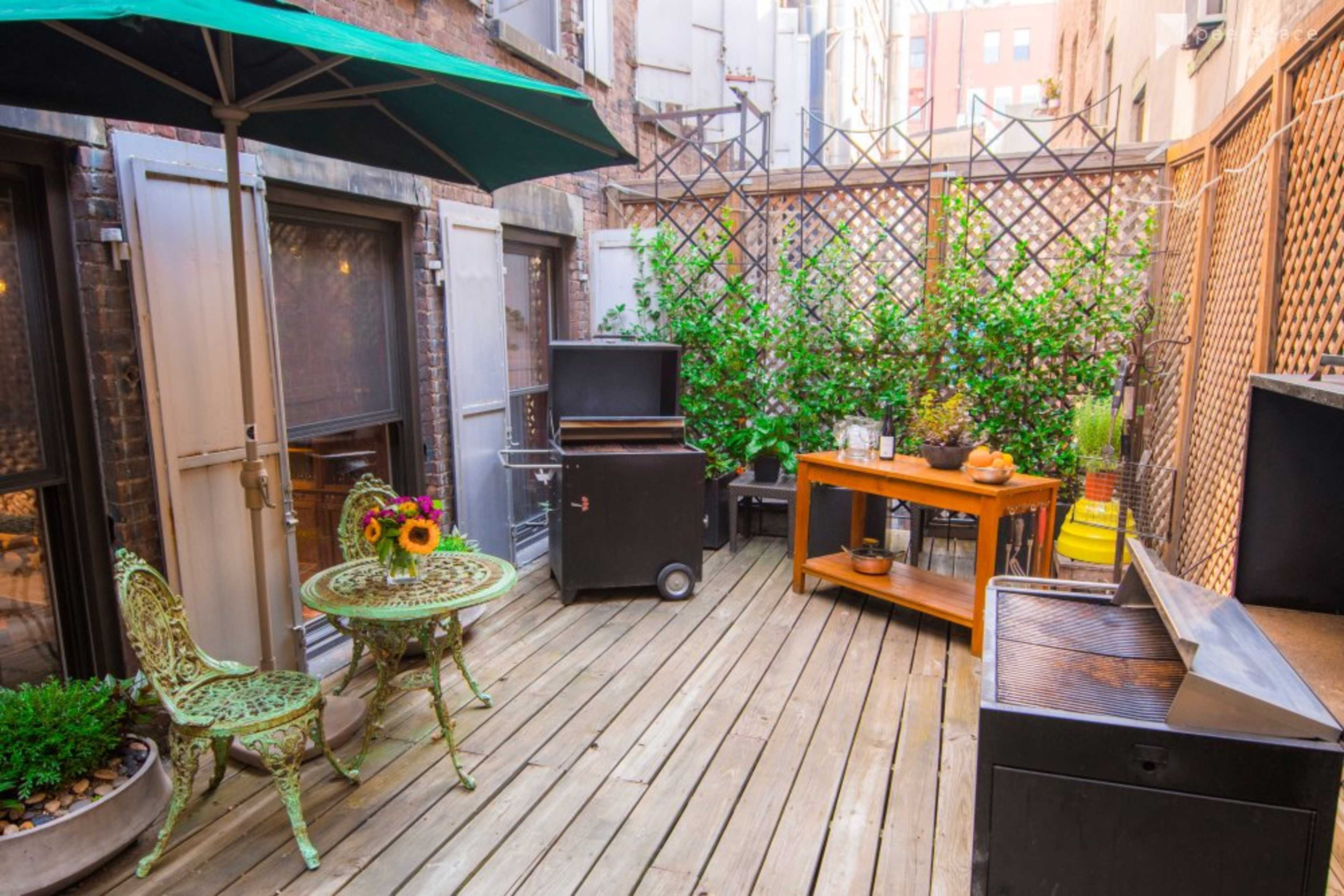 The image shows a small outdoor patio area with a wooden deck, a green umbrella over a table and chairs, and a barbecue grill beside a wooden serving cart.