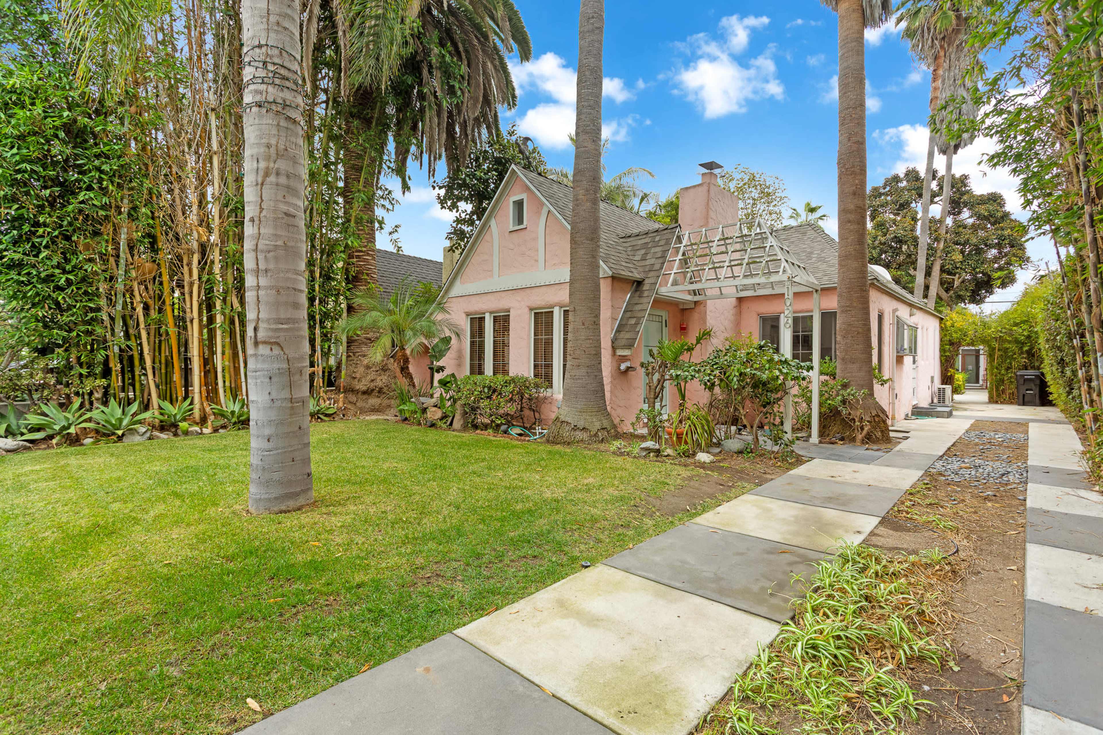 A pink bungalow is surrounded by lush greenery and palm trees, with a stone pathway leading to the entrance.