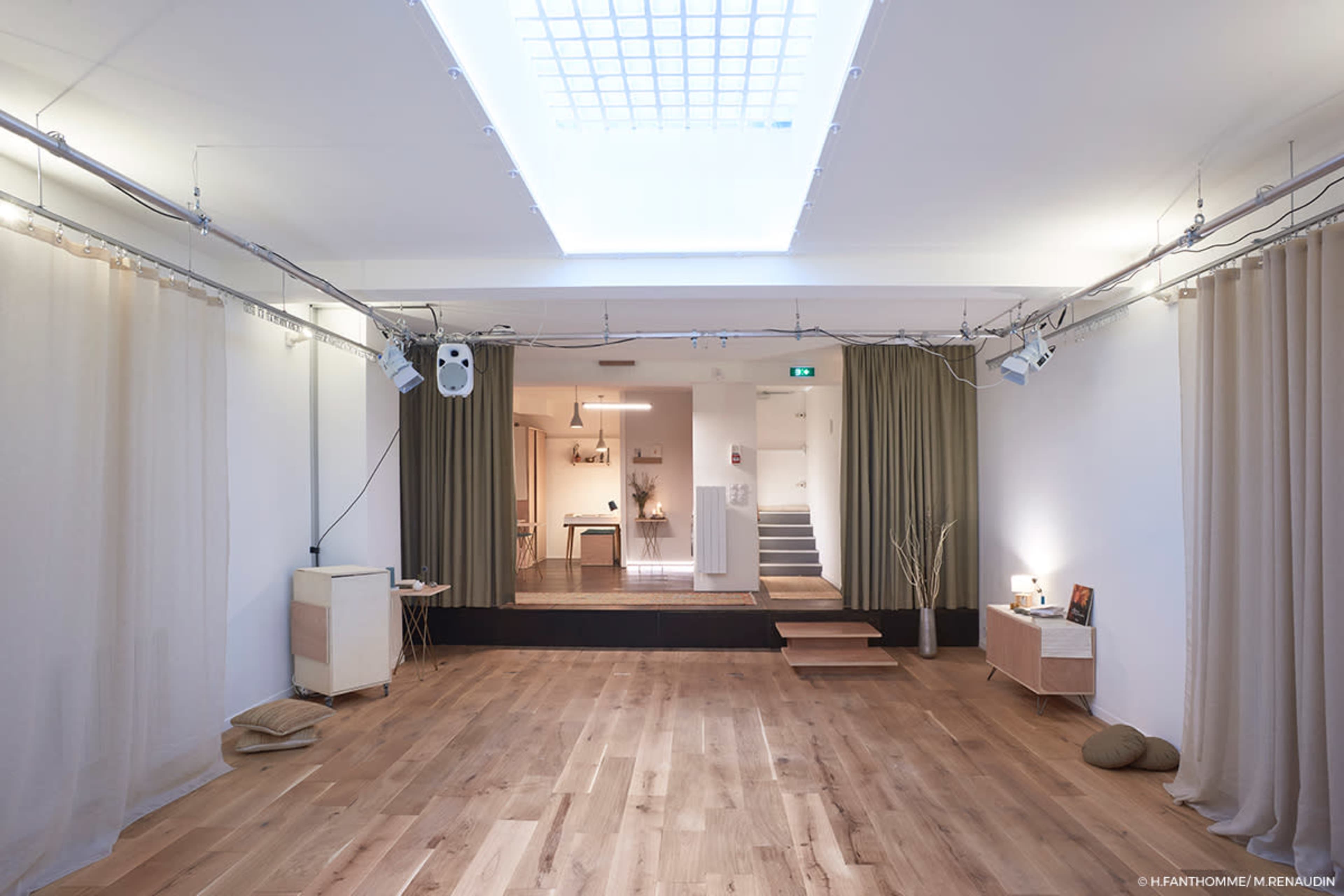 The image shows a minimalist interior space with wooden flooring, cream-colored curtains, and a small staircase leading to an area with a kitchen in the background.