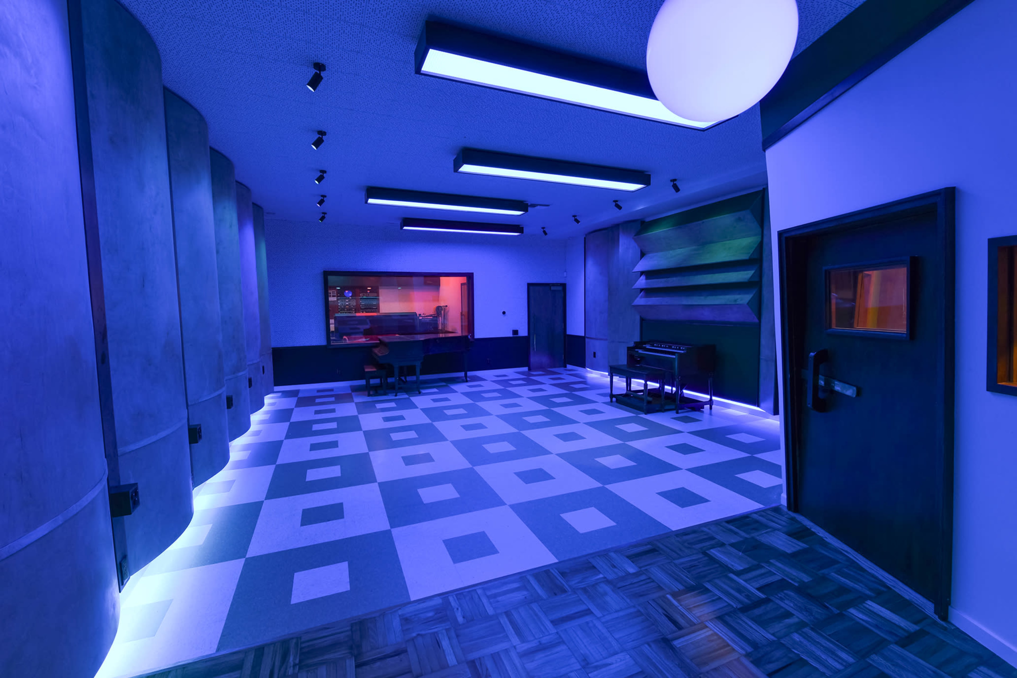 The image shows a modern music studio with blue and purple lighting, featuring a desk, a piano, and soundproof walls.