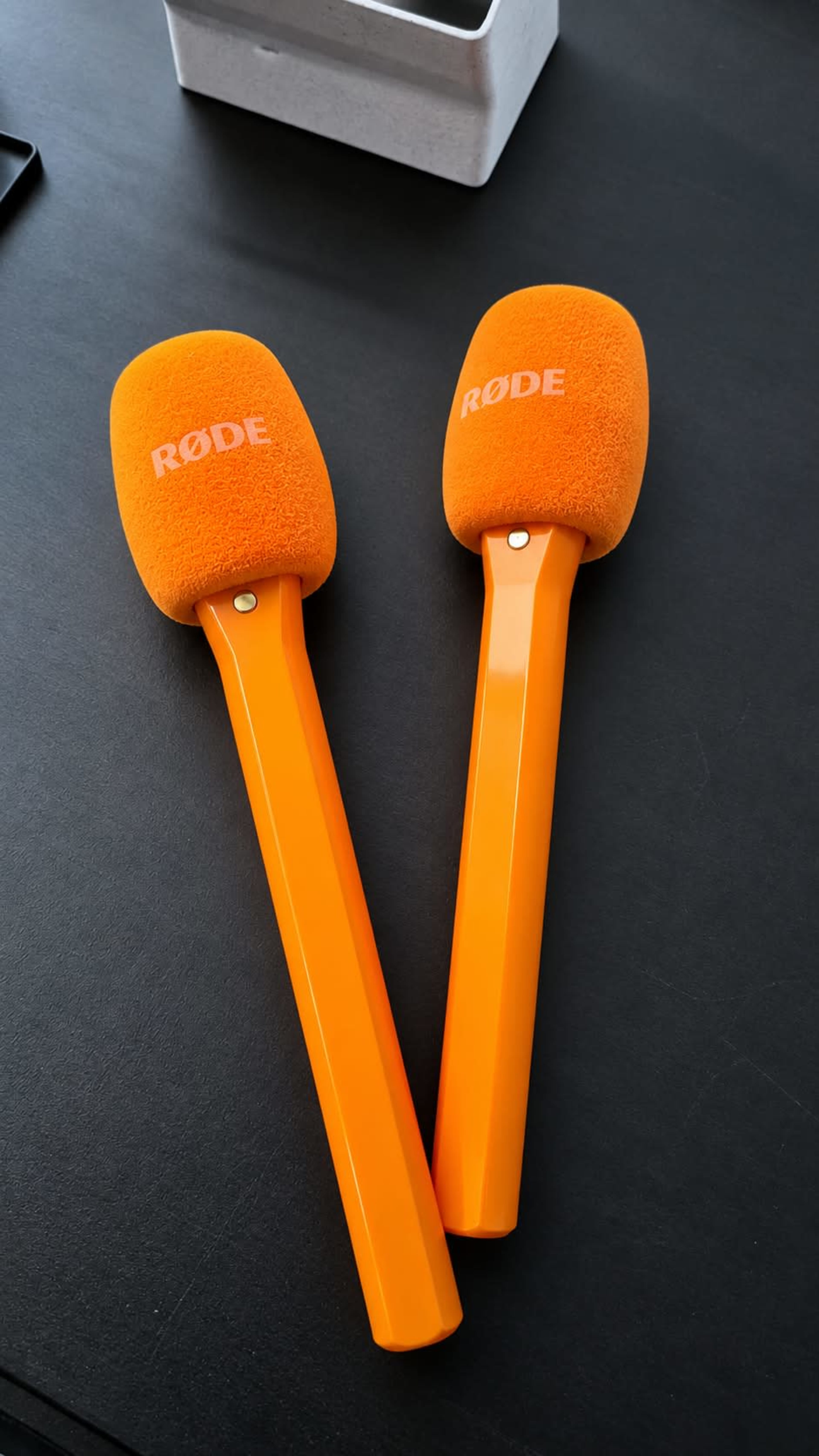 Two orange microphones with a fuzzy cover and a long handle are placed on a black surface.