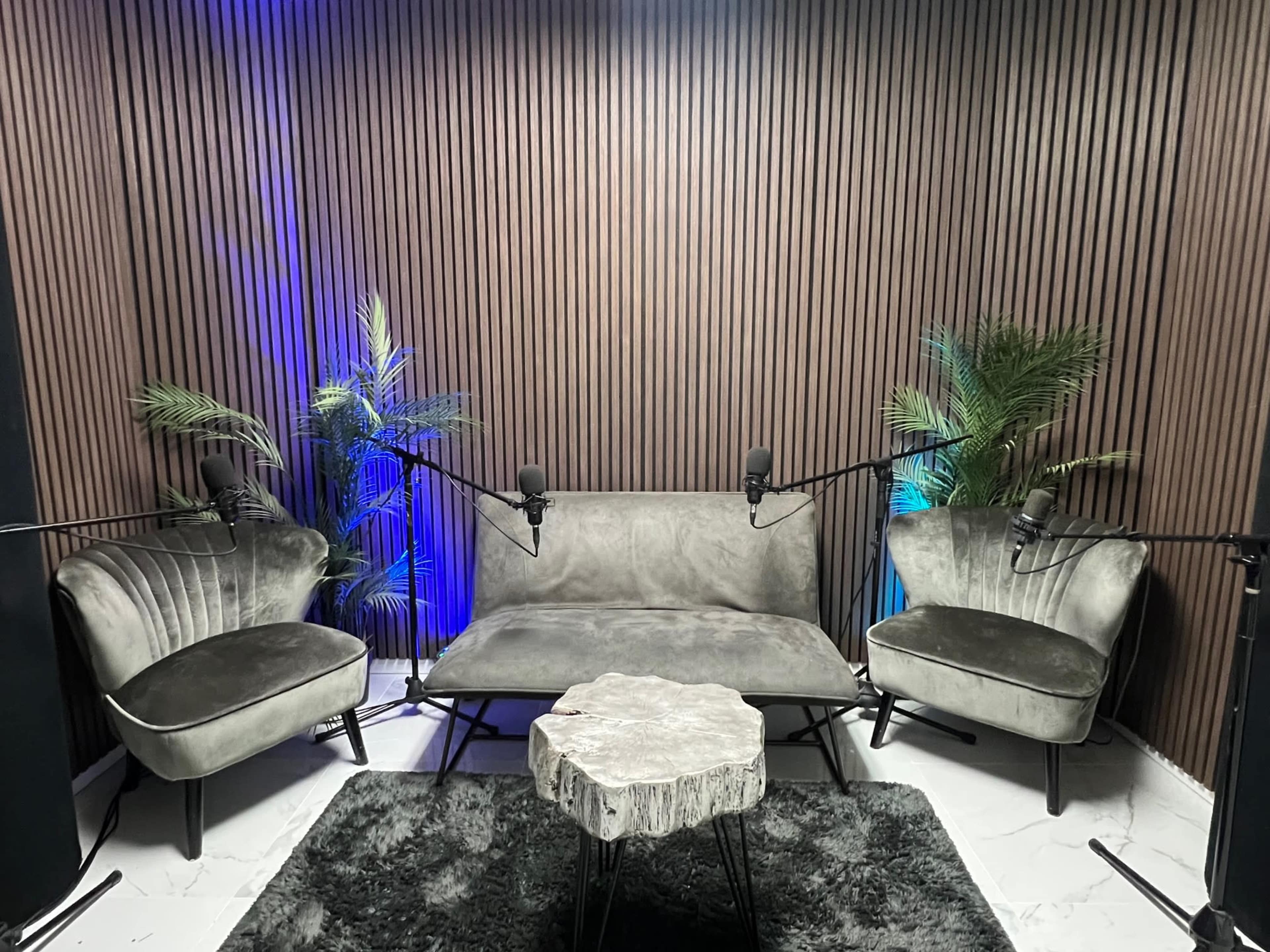 The image shows a modern podcasting studio with two velvet chairs and a sofa, surrounded by plants, all positioned around a small, round table, with microphones set up for recording.