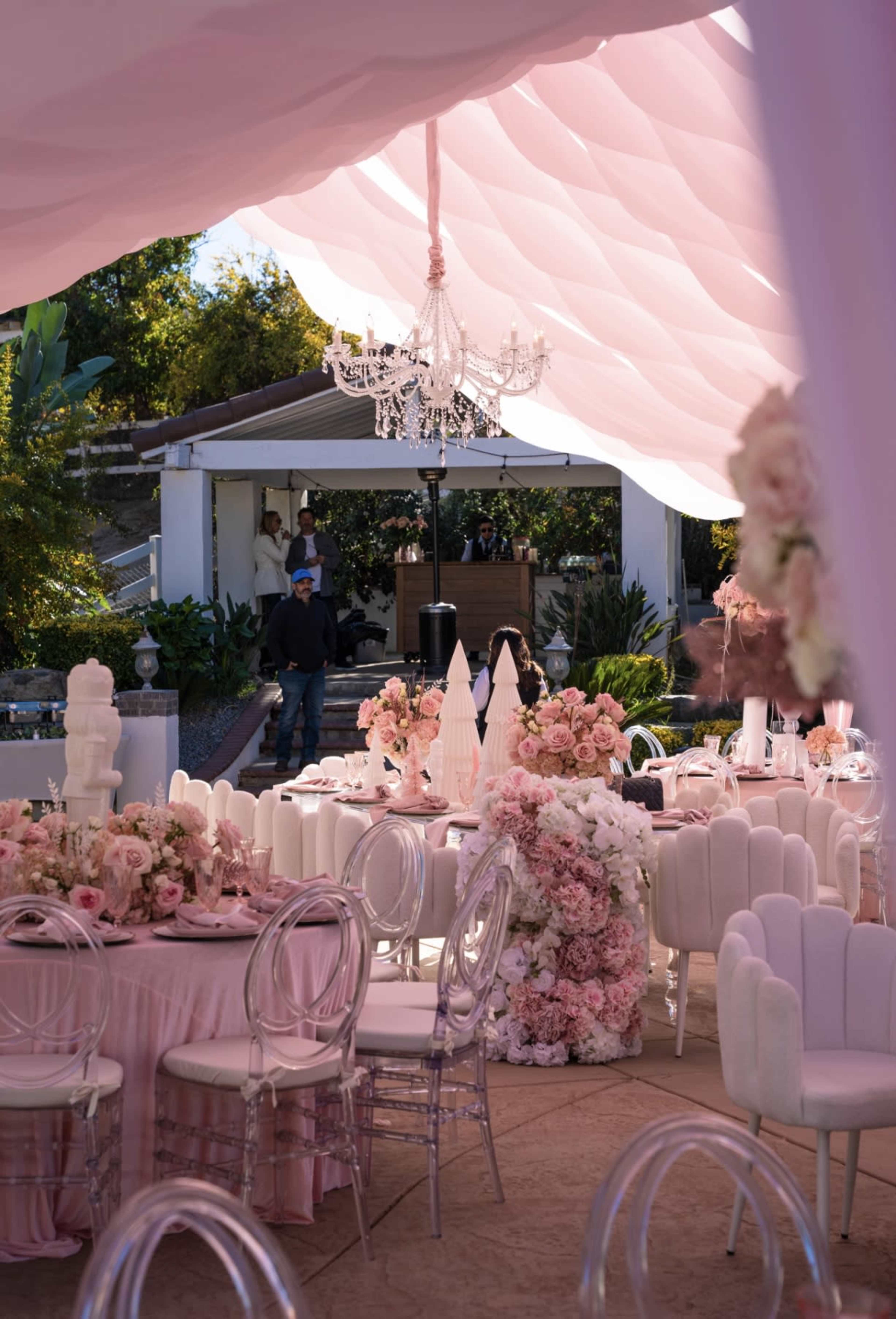 The scene features a beautifully arranged outdoor event space with pink drapery, elegant tables adorned with floral centerpieces, and a chandelier hanging above.