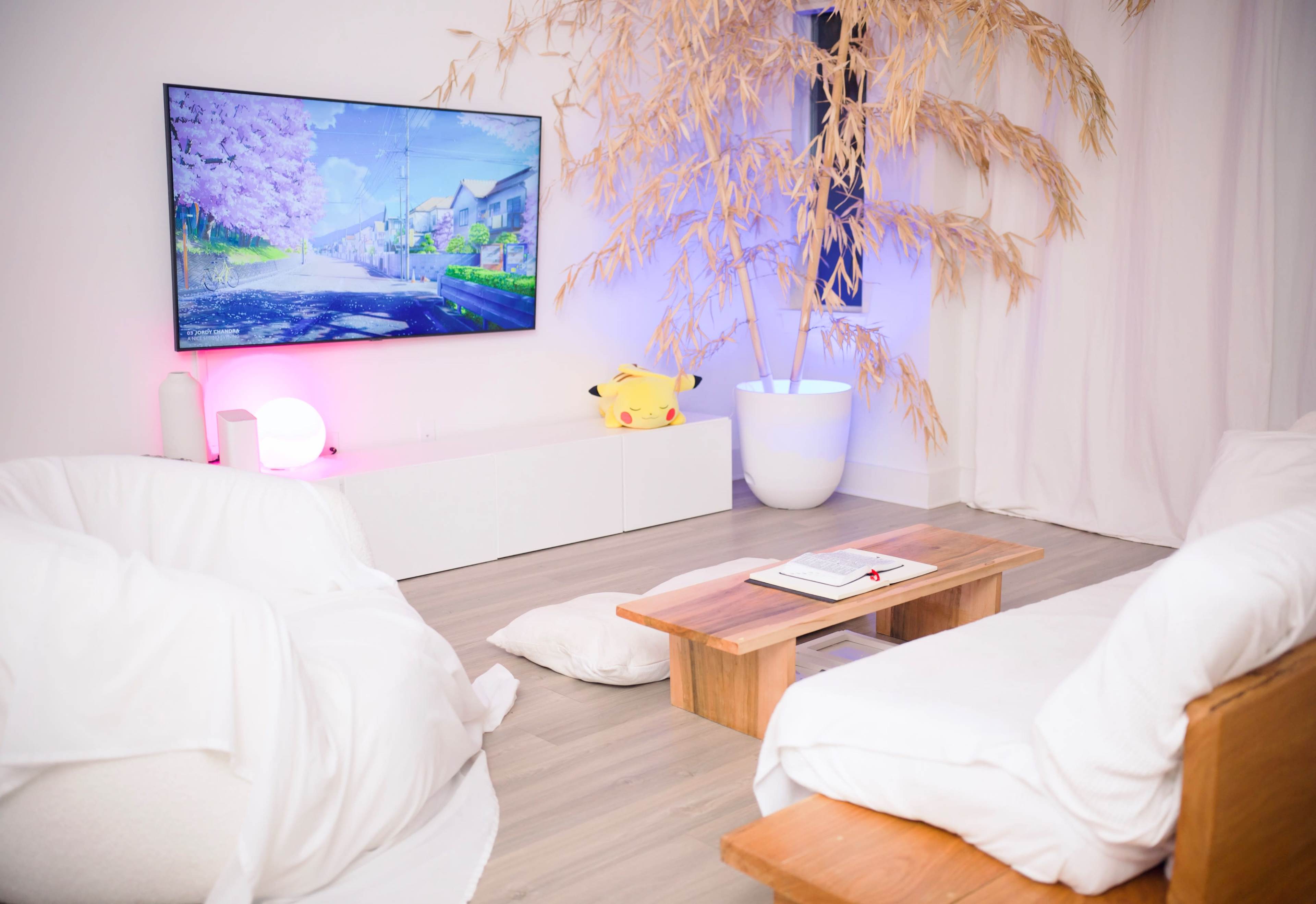 A modern living room features two low wooden coffee tables, white floor cushions, a large wall-mounted television displaying a colorful landscape, and a tall planter with bamboo.