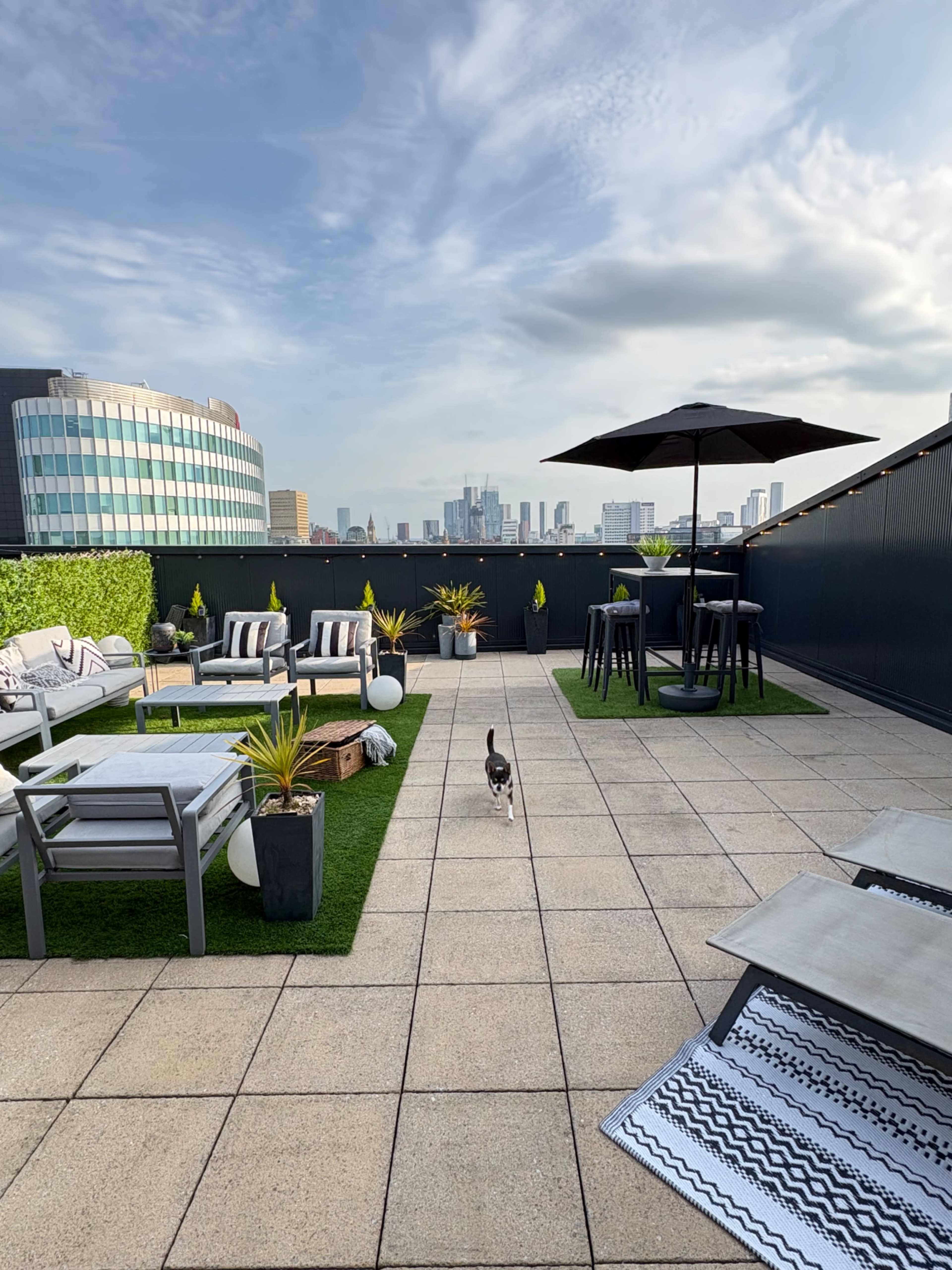 A rooftop terrace features modern seating arrangements, green planters, and a dining table with an umbrella, overlooking a city skyline.