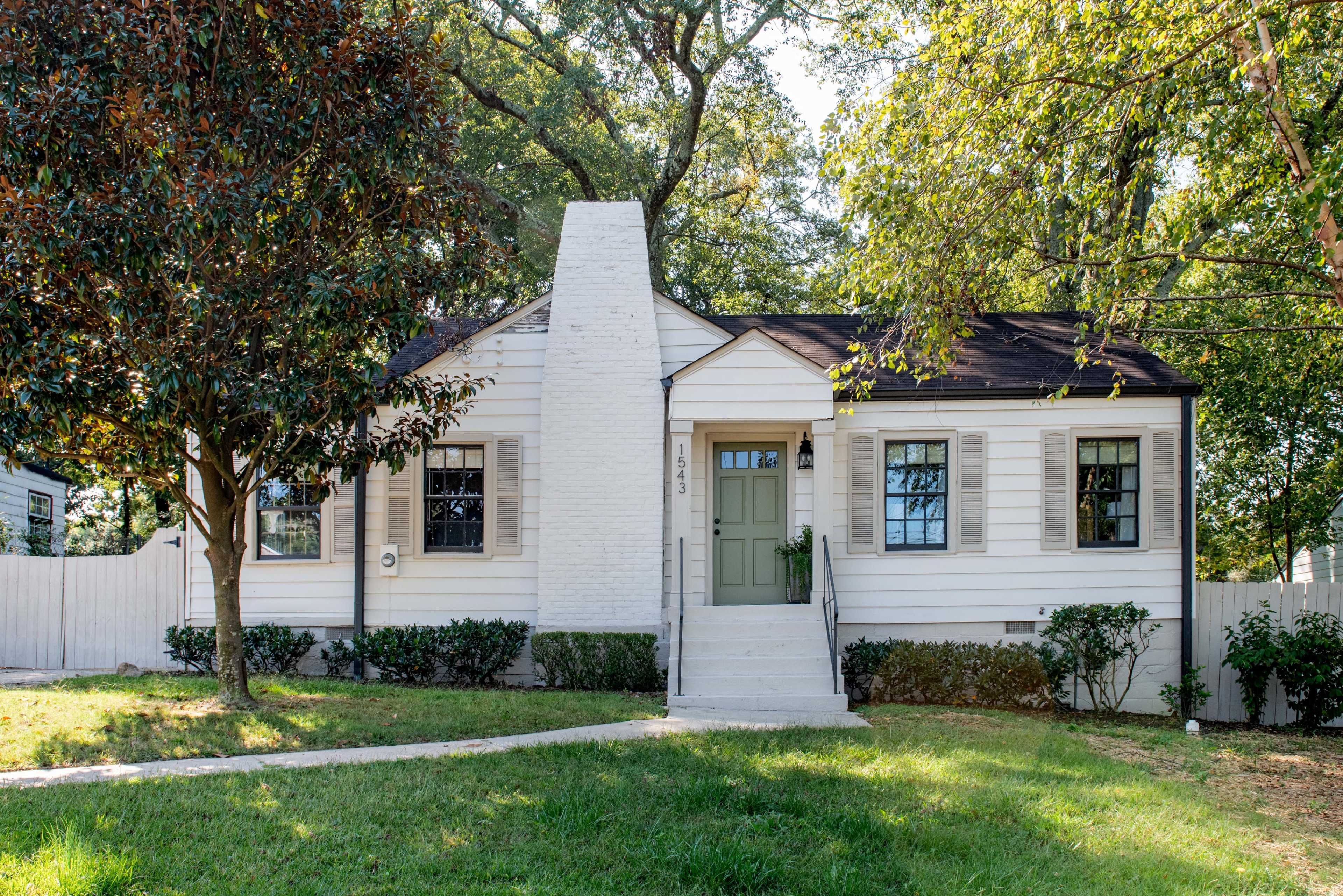 A one-story white cottage with a green door and a central chimney is framed by trees and a manicured lawn.