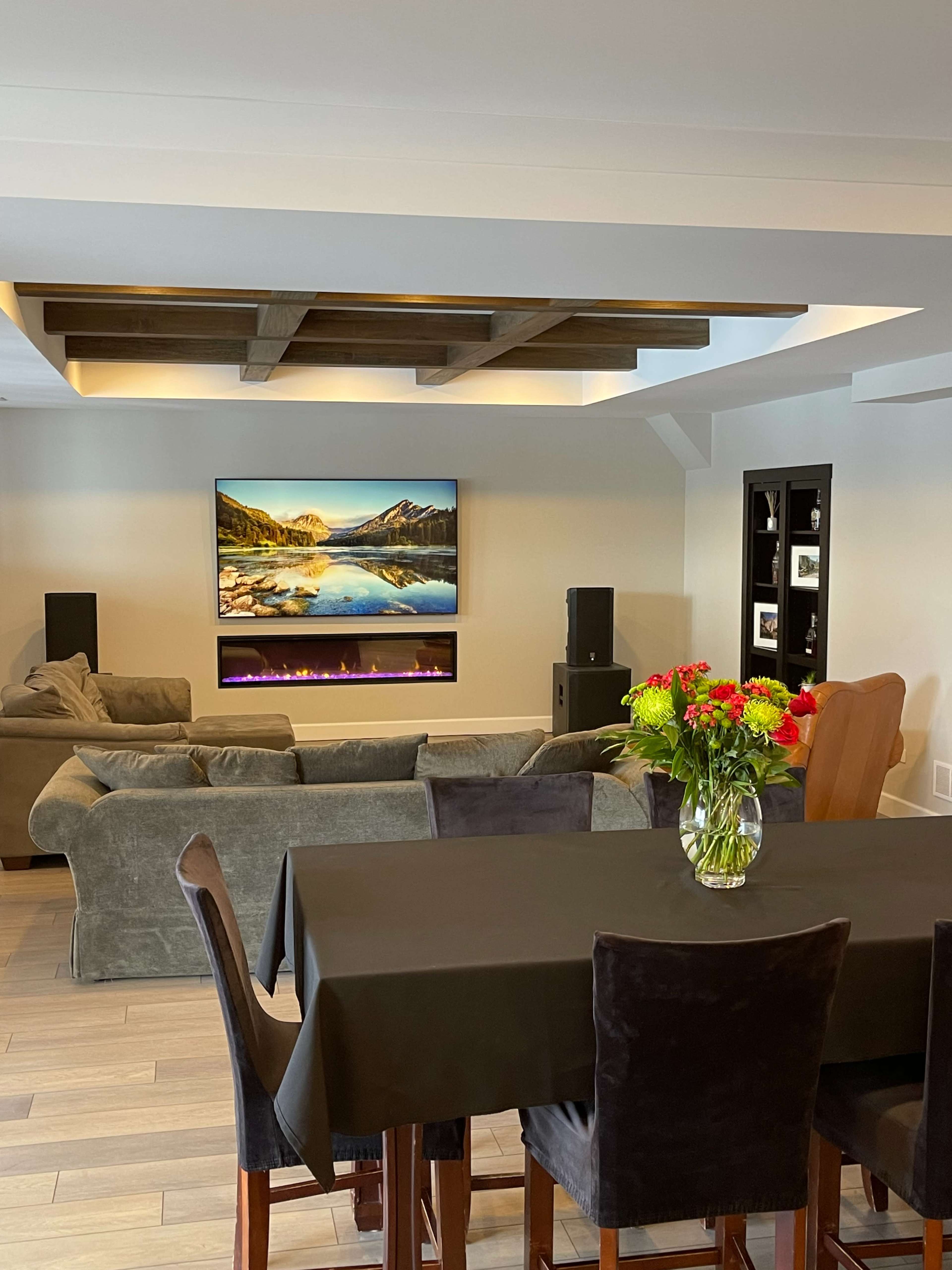 A modern living room with a dark dining table, a sectional sofa, an entertainment center featuring a flat-screen TV above a linear fireplace, and a vase of flowers on the table.