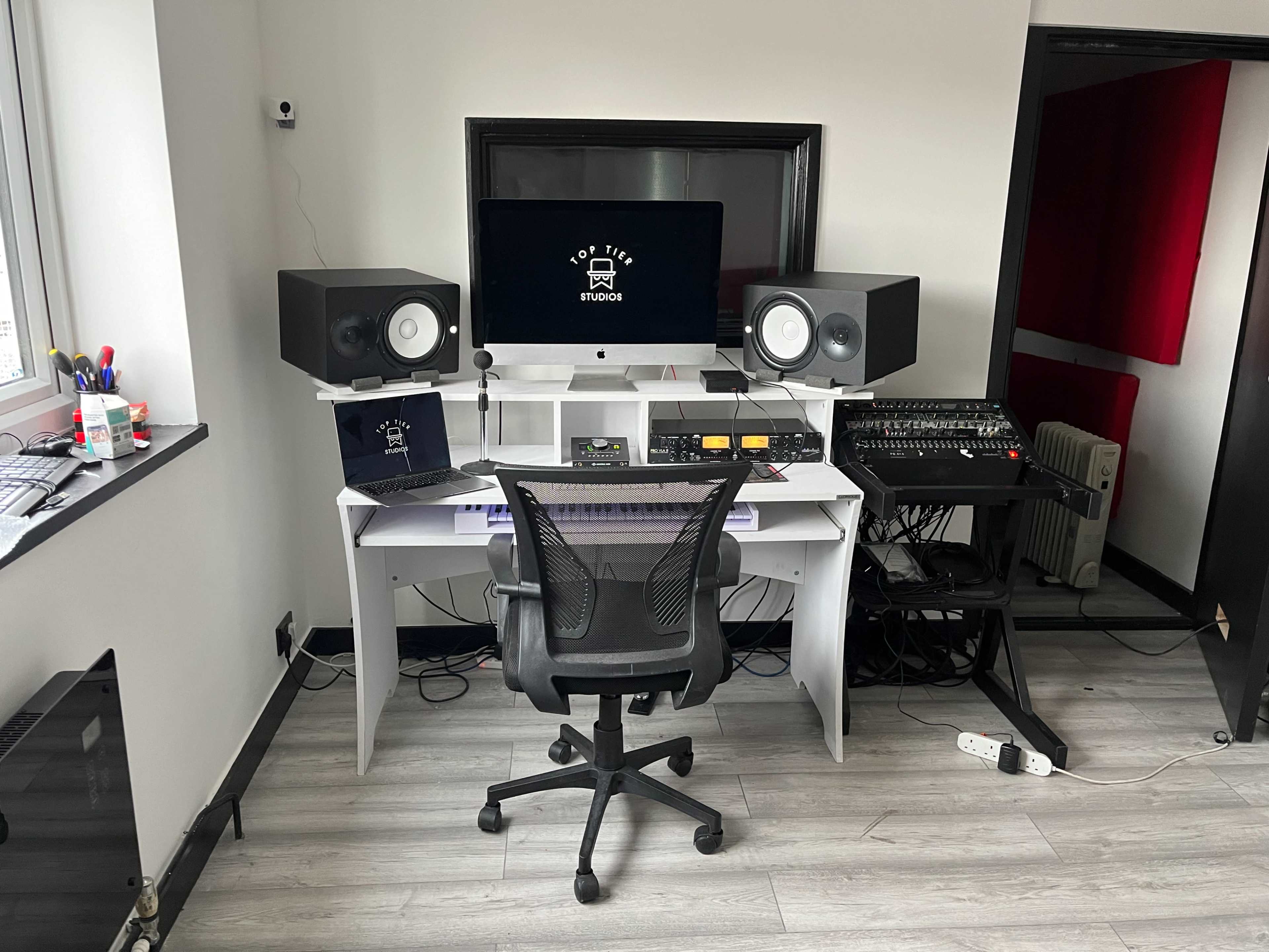 A modern music studio features a desk with computer monitors, audio equipment, and studio monitors positioned in a well-lit room.