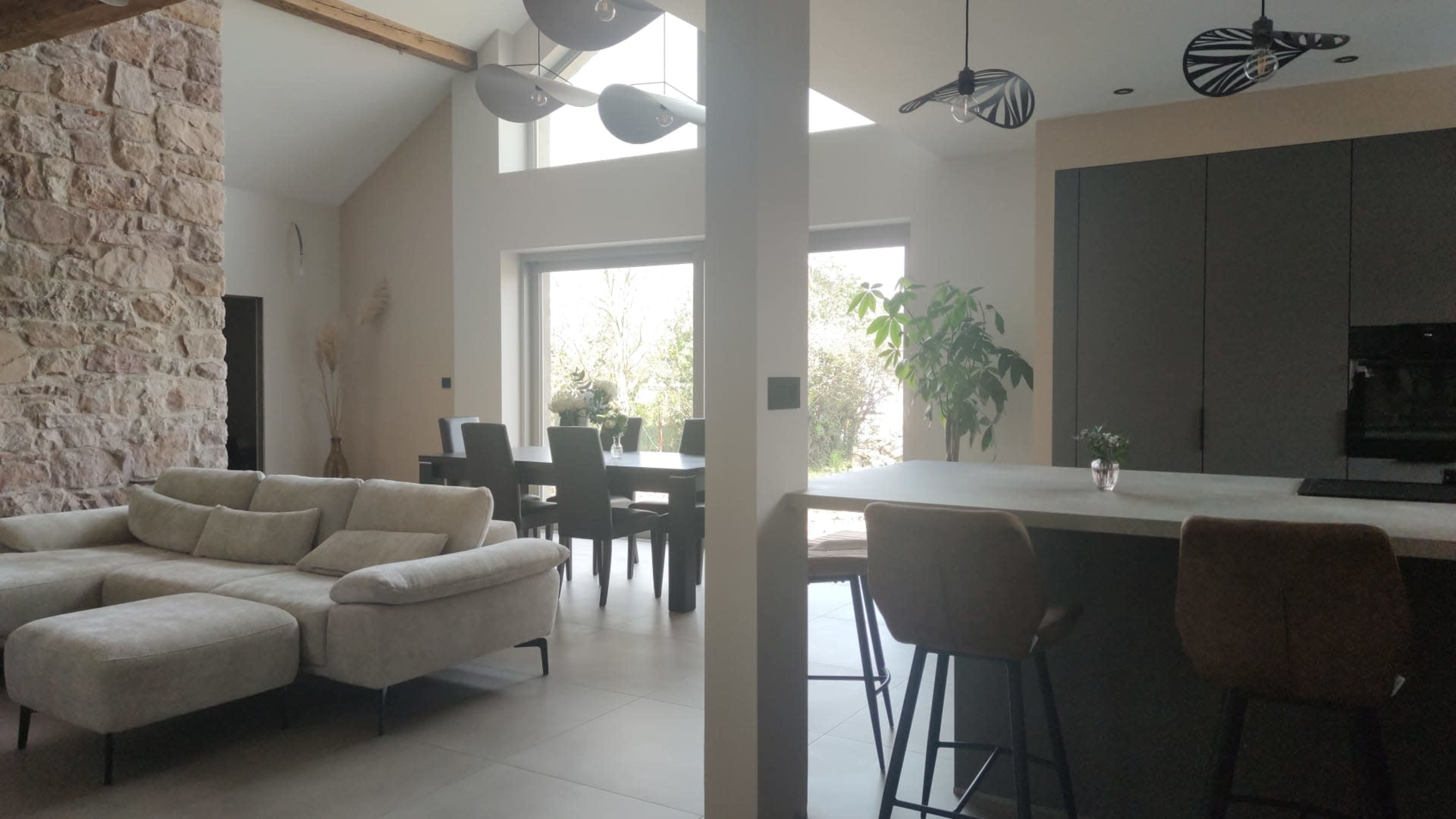 The image shows a modern open-concept living space featuring a light-colored sectional sofa, a dining area with a black dining table and chairs, and a kitchen with bar stools and dark cabinetry.