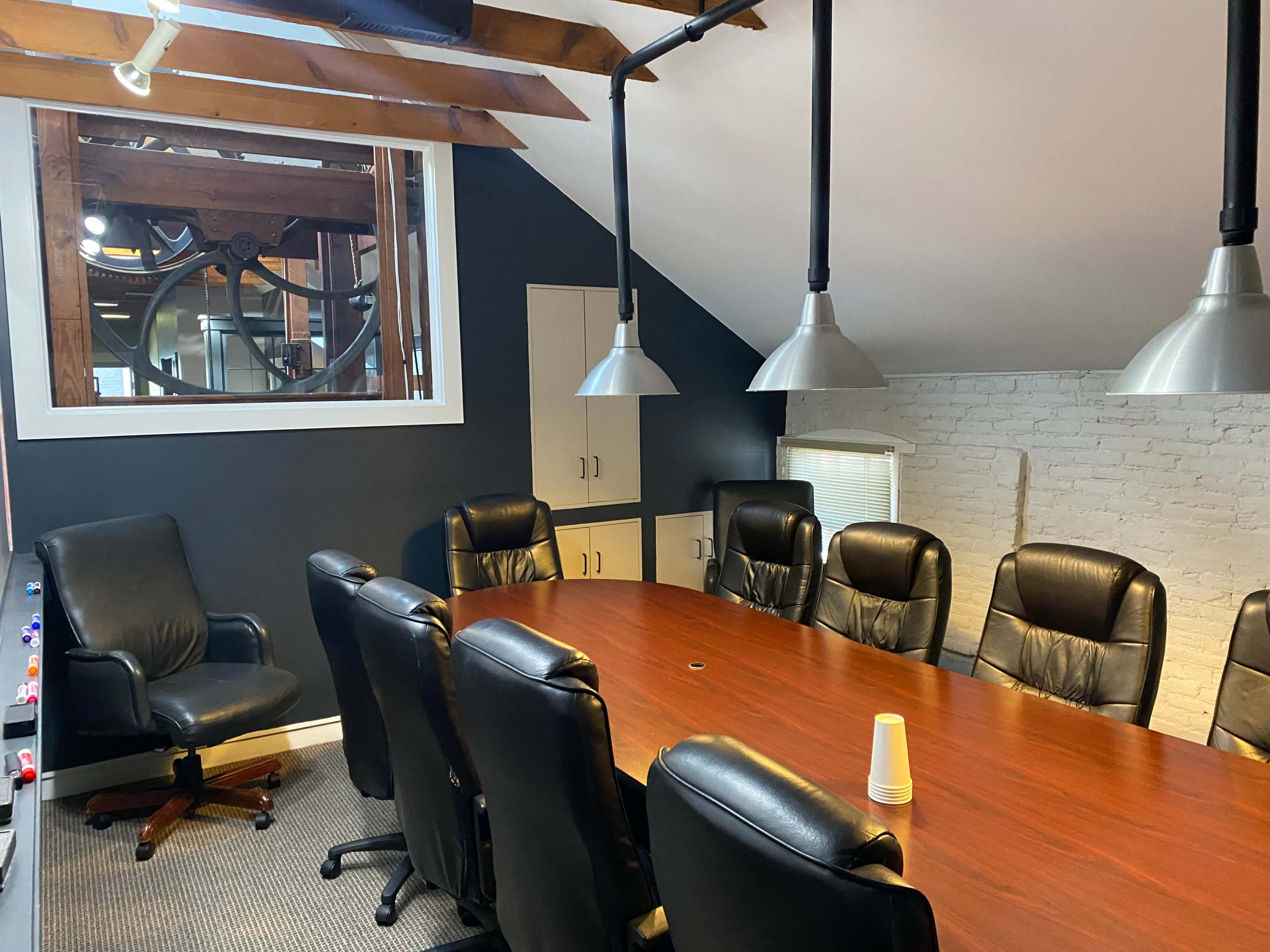 The image shows a conference room with a long wooden table surrounded by black leather chairs and large industrial-style ceiling lights.