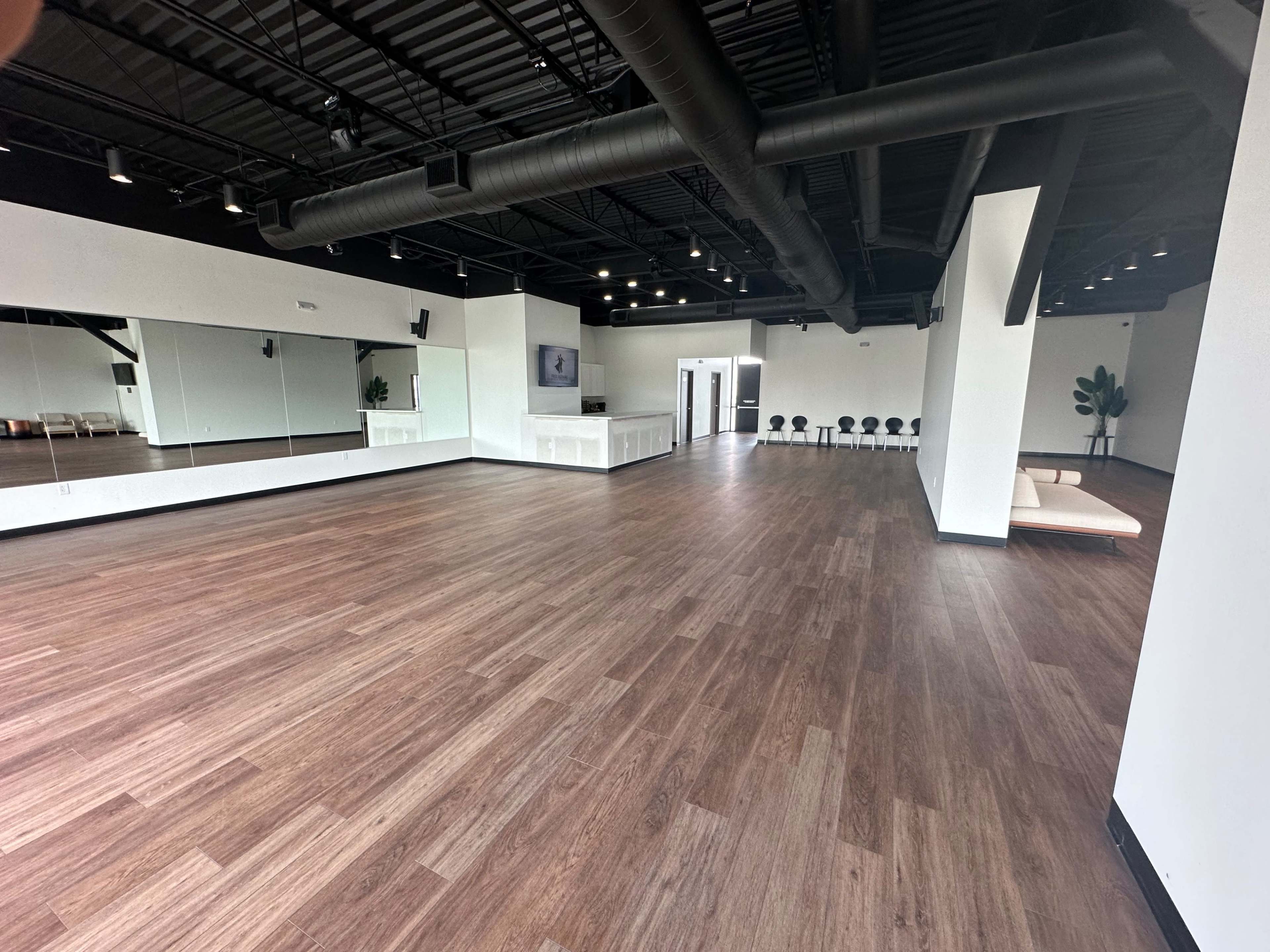 The image shows a spacious, well-lit studio with wooden flooring, large mirrors on one wall, and a seating area in the corner.