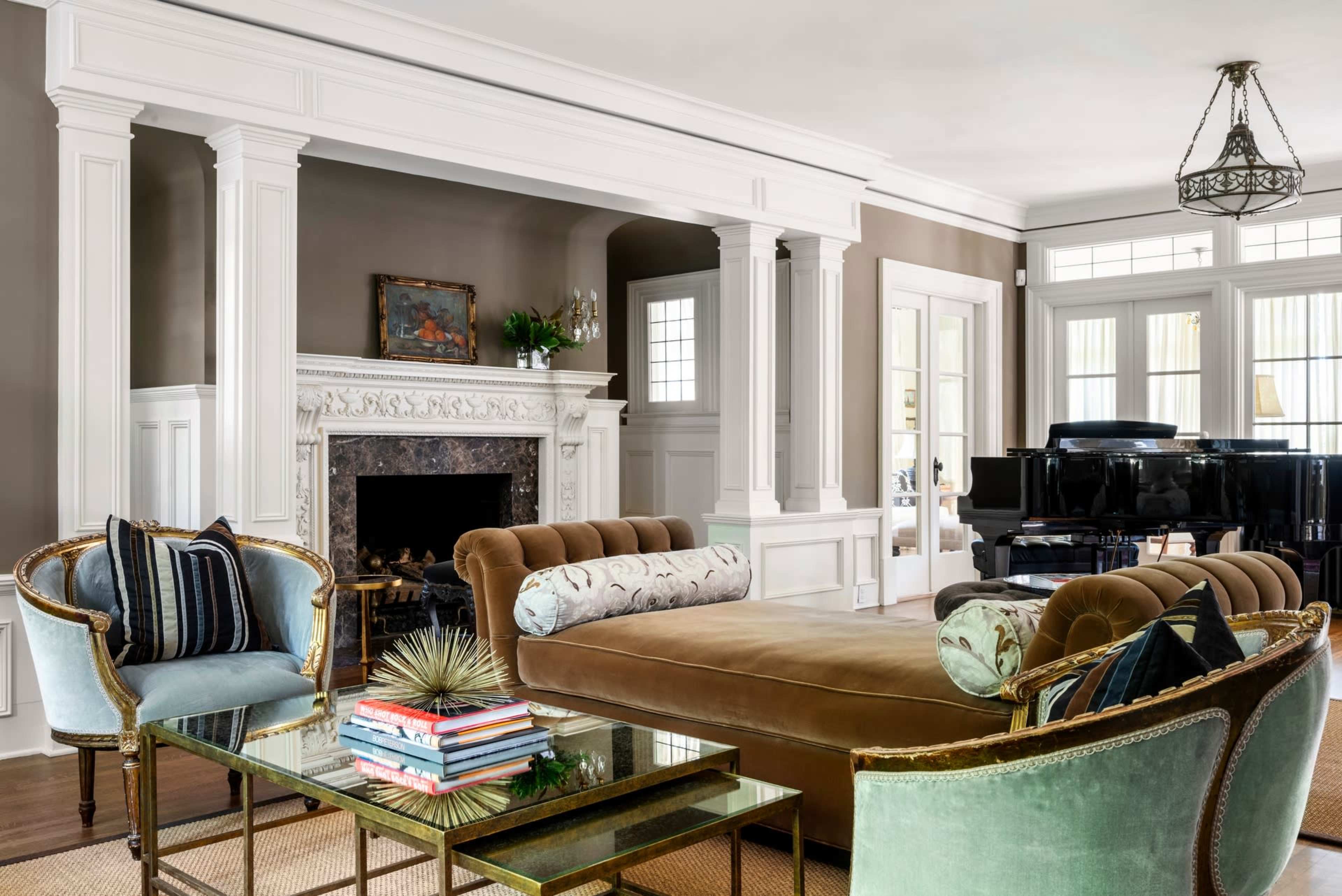 A living room with a velvet chaise lounge, a glass coffee table stacked with books, a grand piano in the background, and a decorative fireplace framed by white columns.