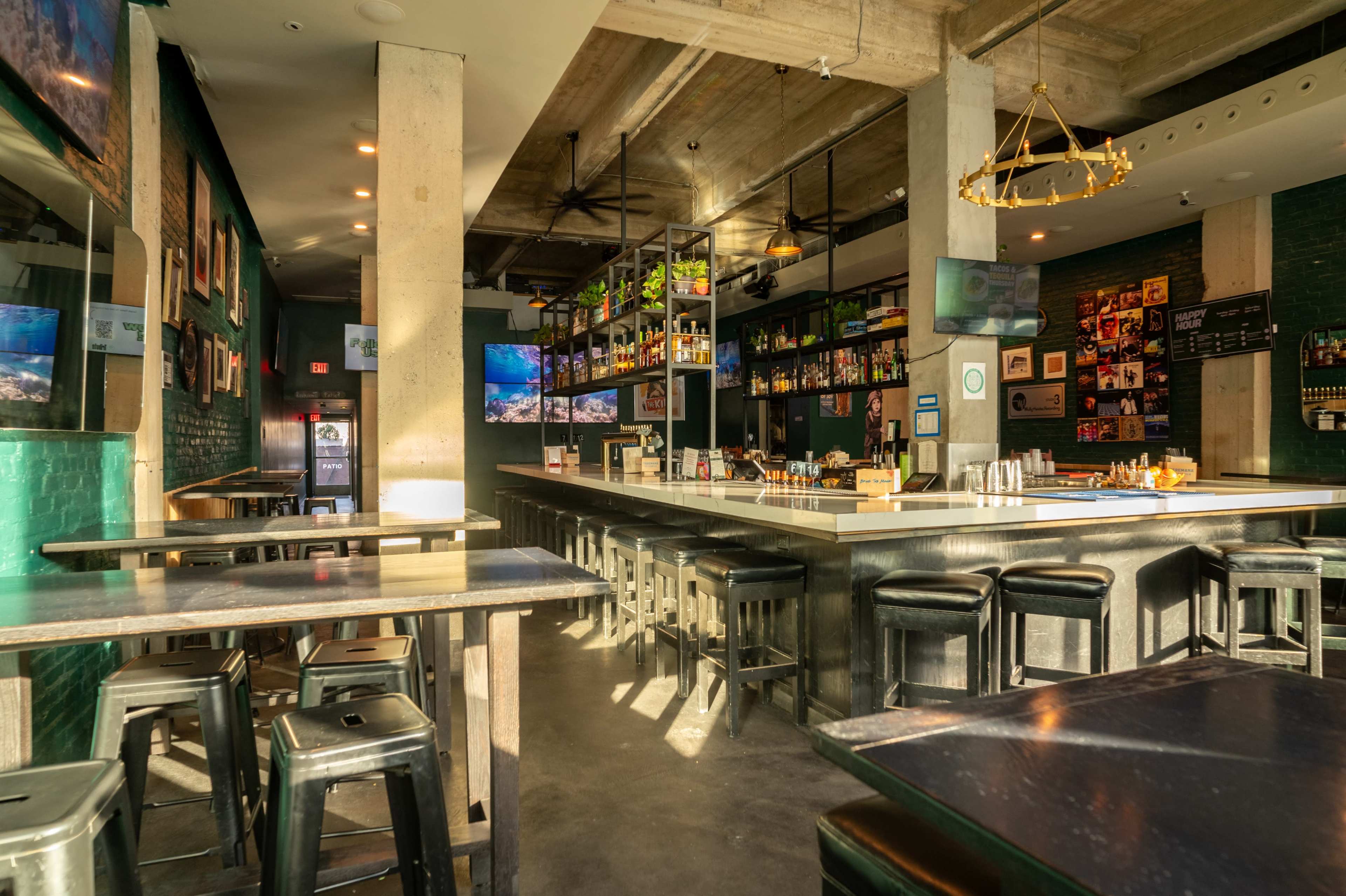 The image shows a modern bar interior with a long countertop, high stools, and shelves lined with bottles against a backdrop of green walls and large windows.