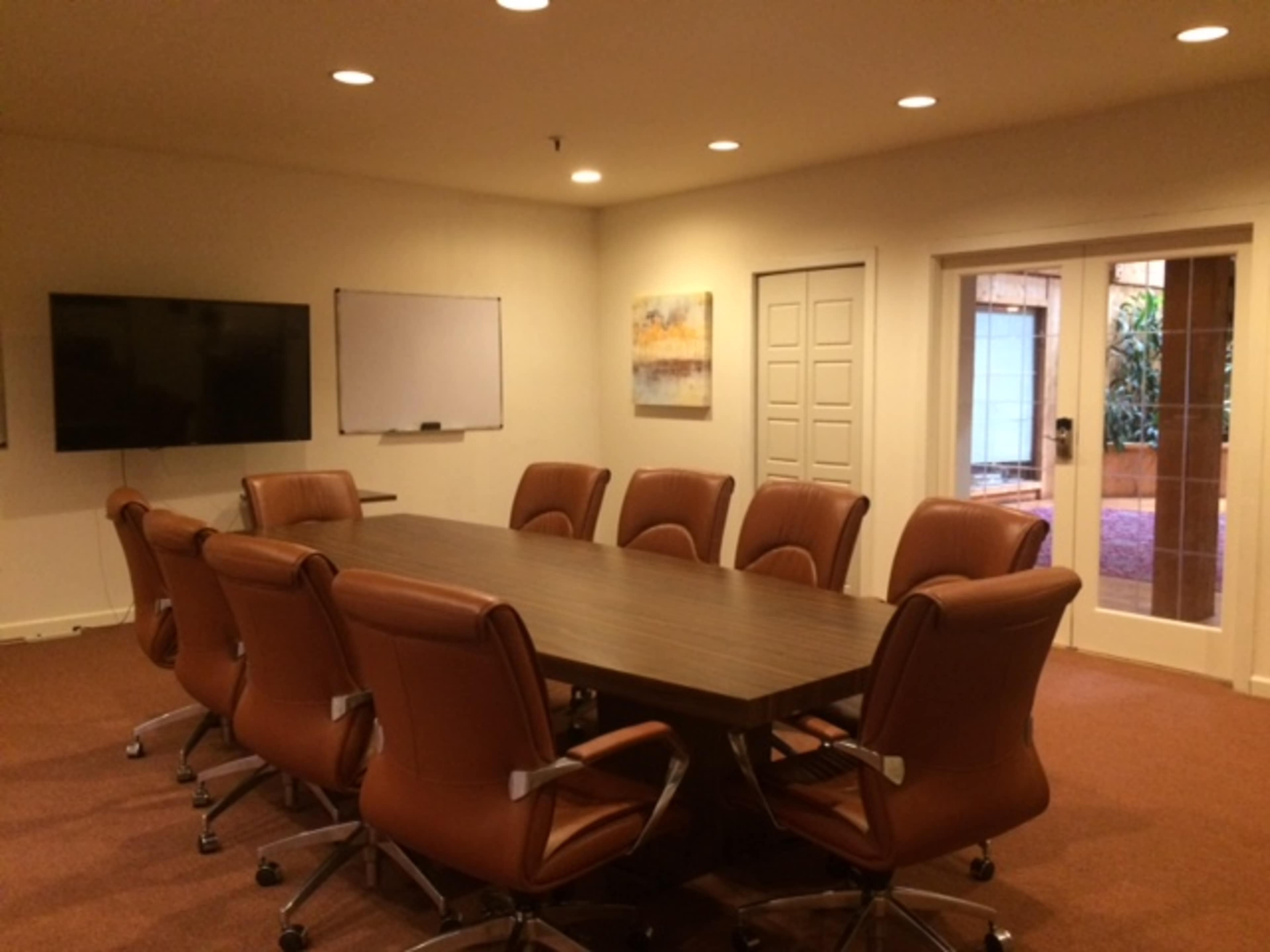 A conference room features a large wooden table surrounded by ten brown leather chairs, with a television and whiteboard on one wall.