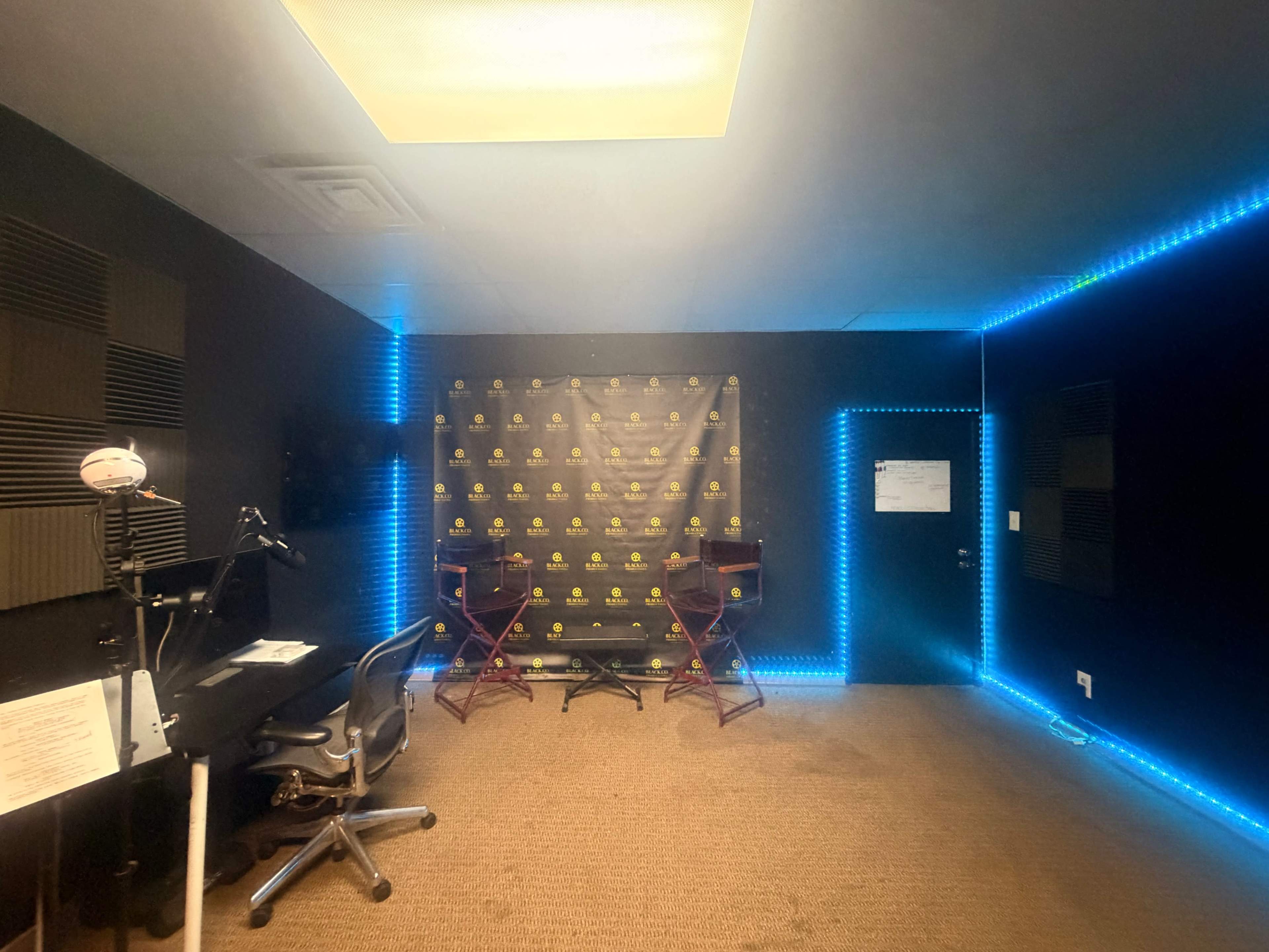 The image shows a sound studio with black walls, stylish lighting, and two director's chairs in front of a backdrop featuring a logo.
