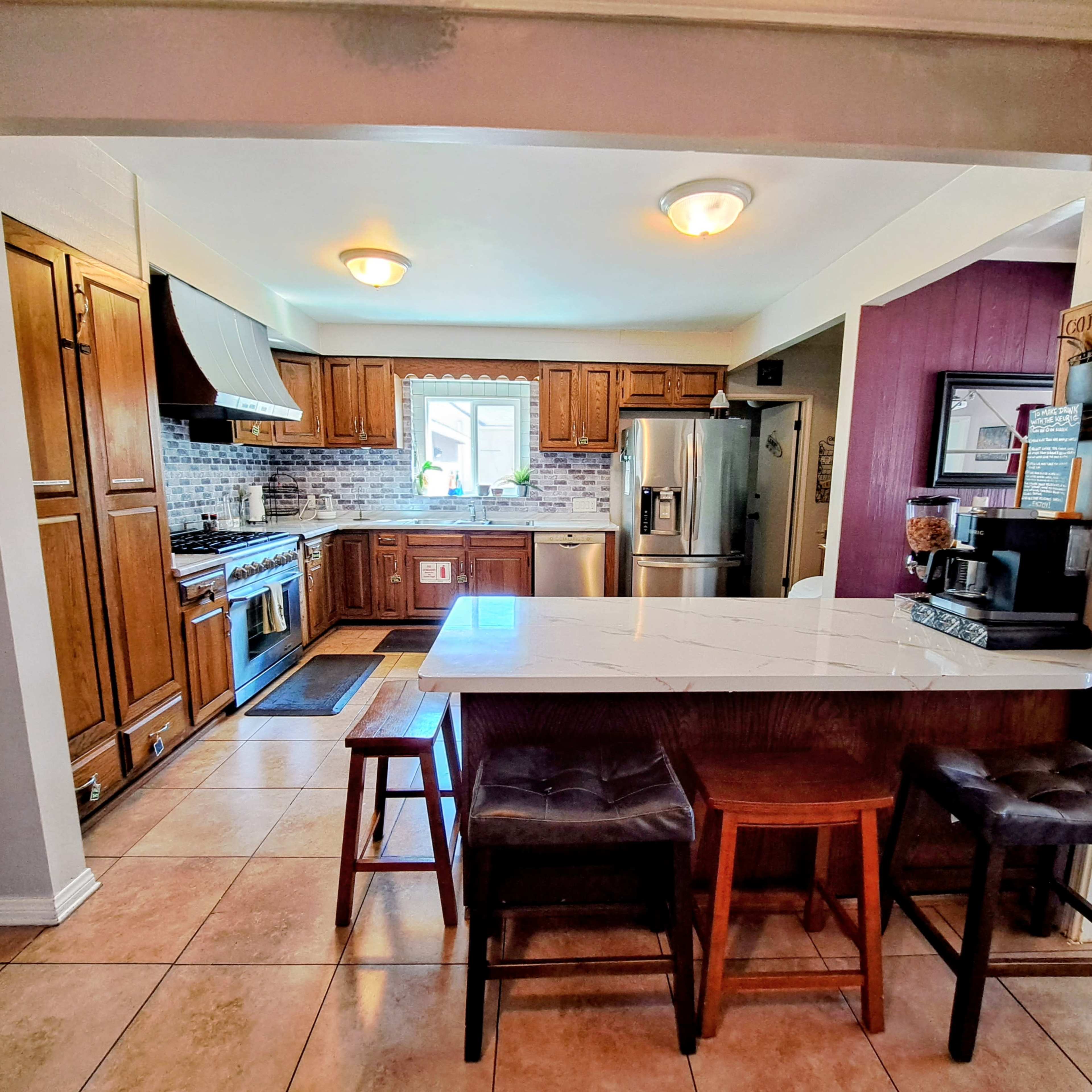 The image shows a modern kitchen featuring wooden cabinets, stainless steel appliances, a tile floor, and a central island with seating.