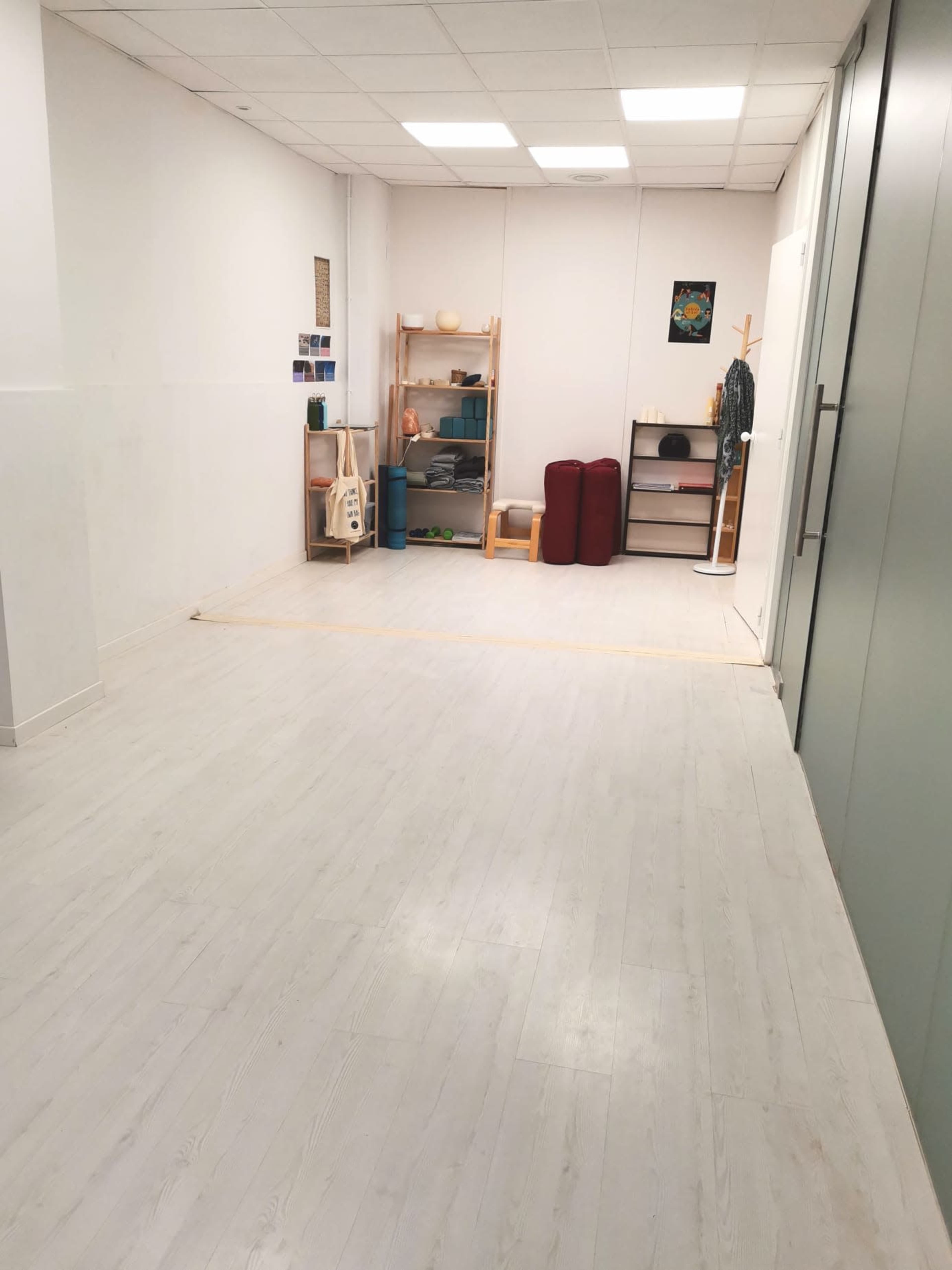 The image shows a minimalist room with light-colored flooring and shelves containing items, including yoga mats and cushions, against a white wall.