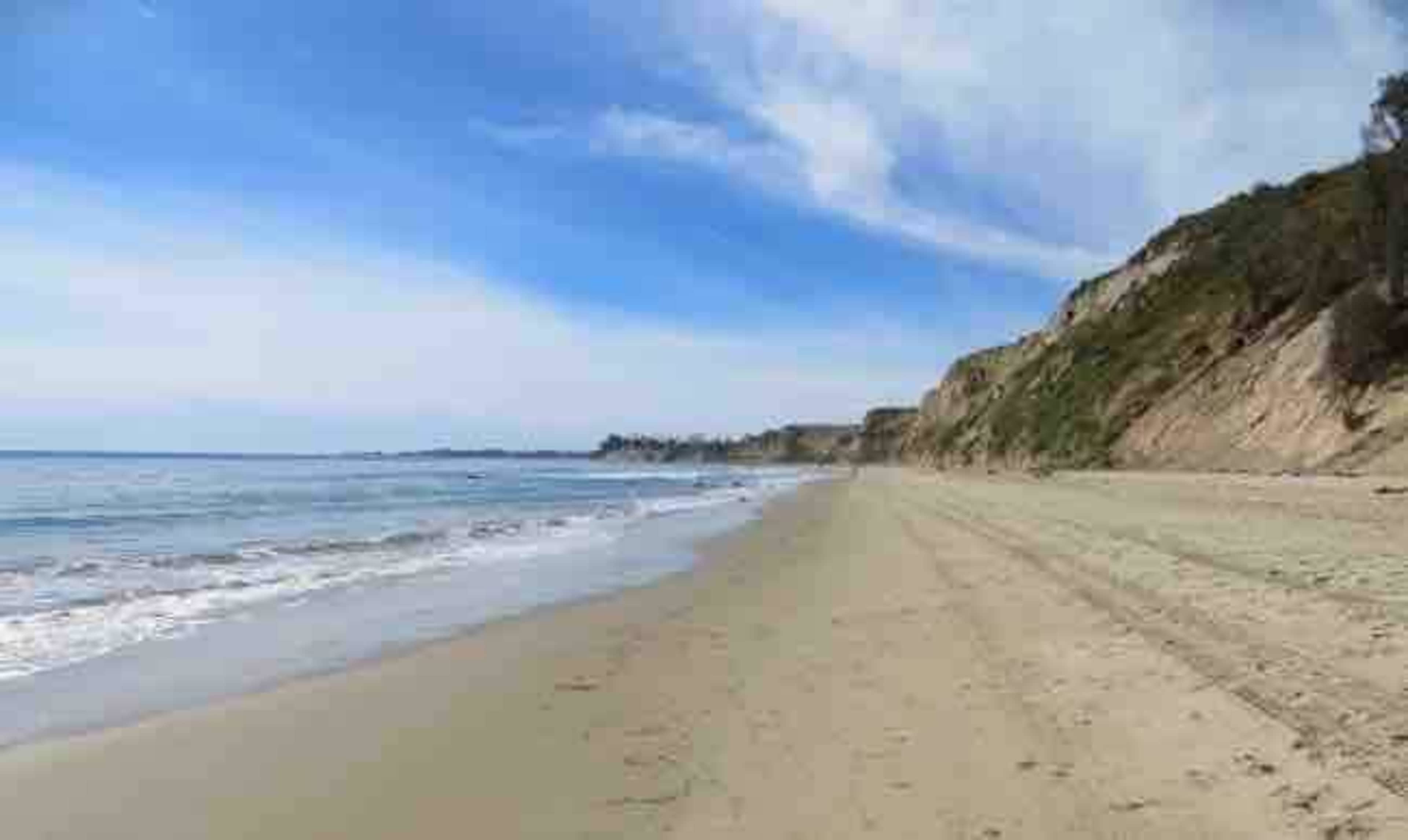 A sandy beach stretches along the shore, with gentle waves lapping at the water's edge and cliffs rising in the background under a blue sky.