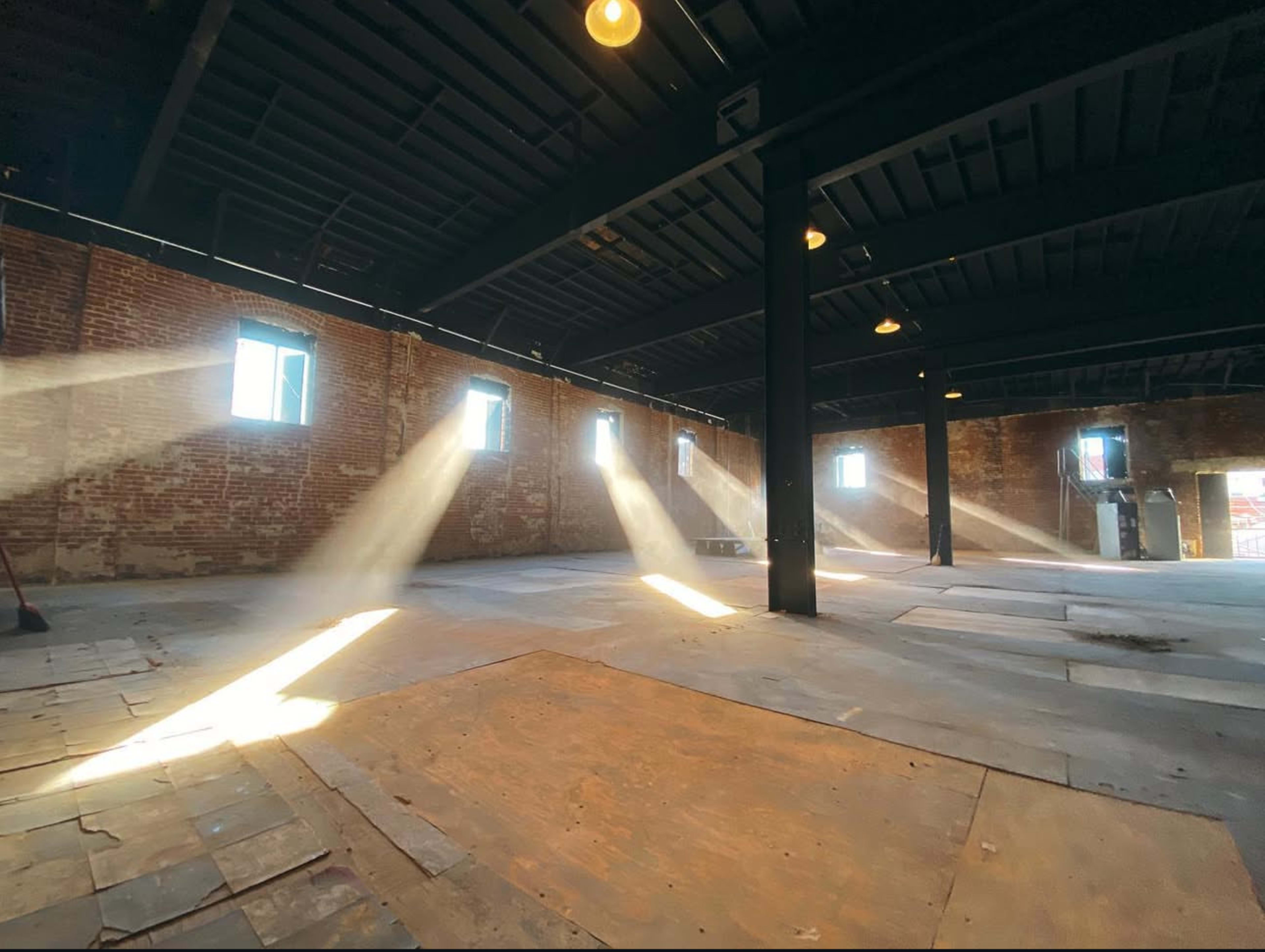 Sunlight streams through windows in an empty industrial space with exposed brick walls and wooden flooring.