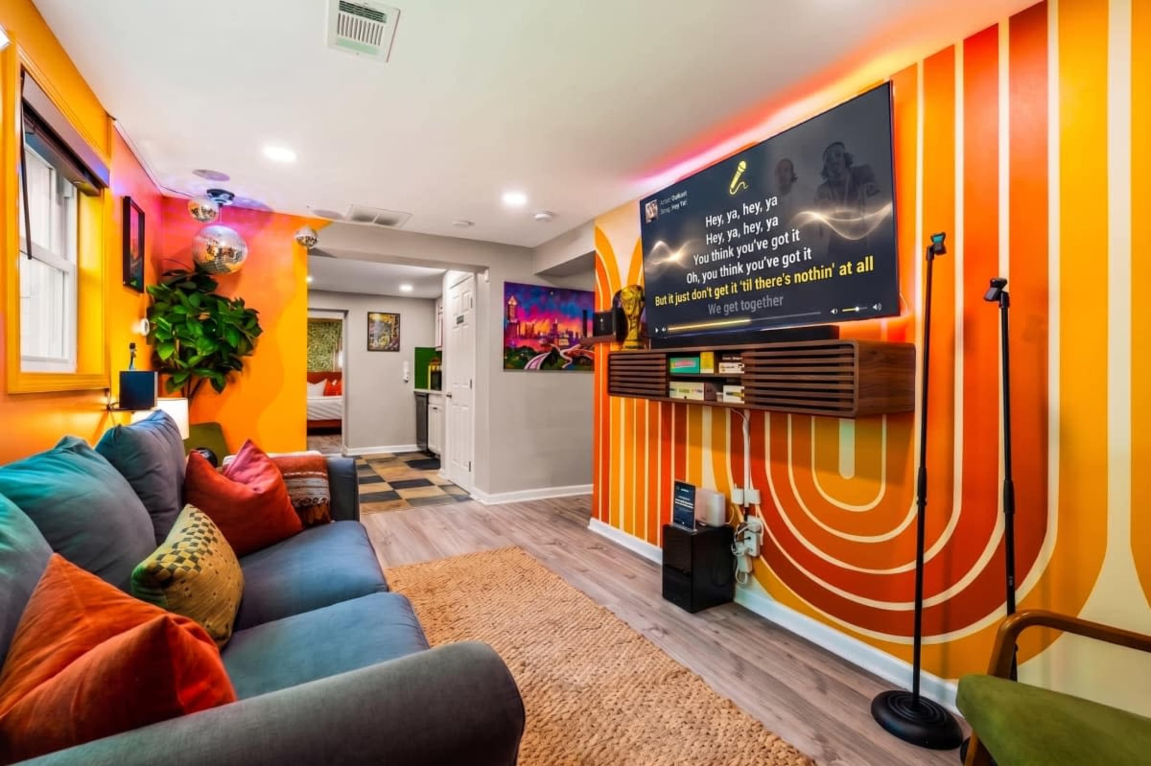 The room features a colorful retro design with an orange-striped wall, a large TV displaying karaoke lyrics, a gray couch, and a woven rug on the floor.
