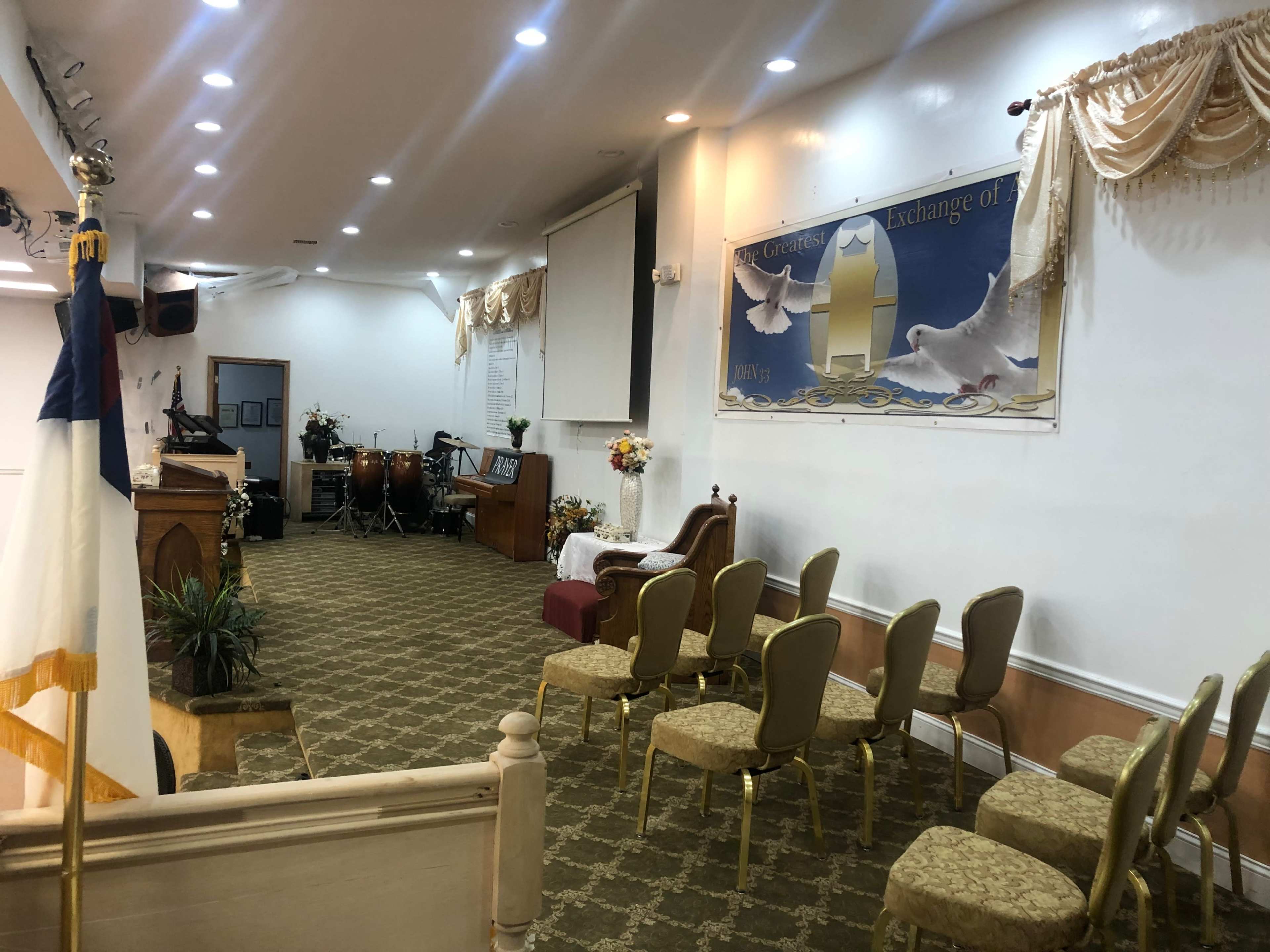 The image shows the interior of a small church with wooden furniture, seating arranged in rows, and a stage area featuring musical instruments and a wall mural with doves.