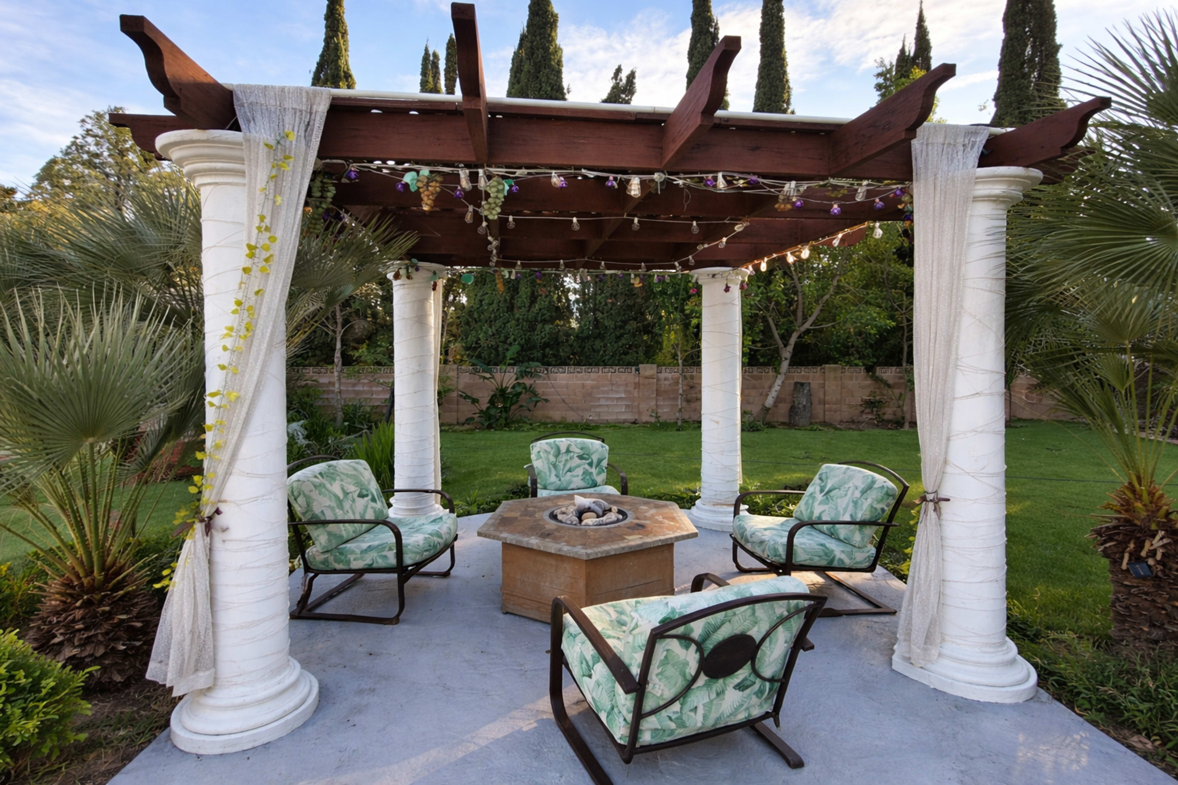 A pergola with four columns and a seating area surrounds a fire pit in a landscaped garden.