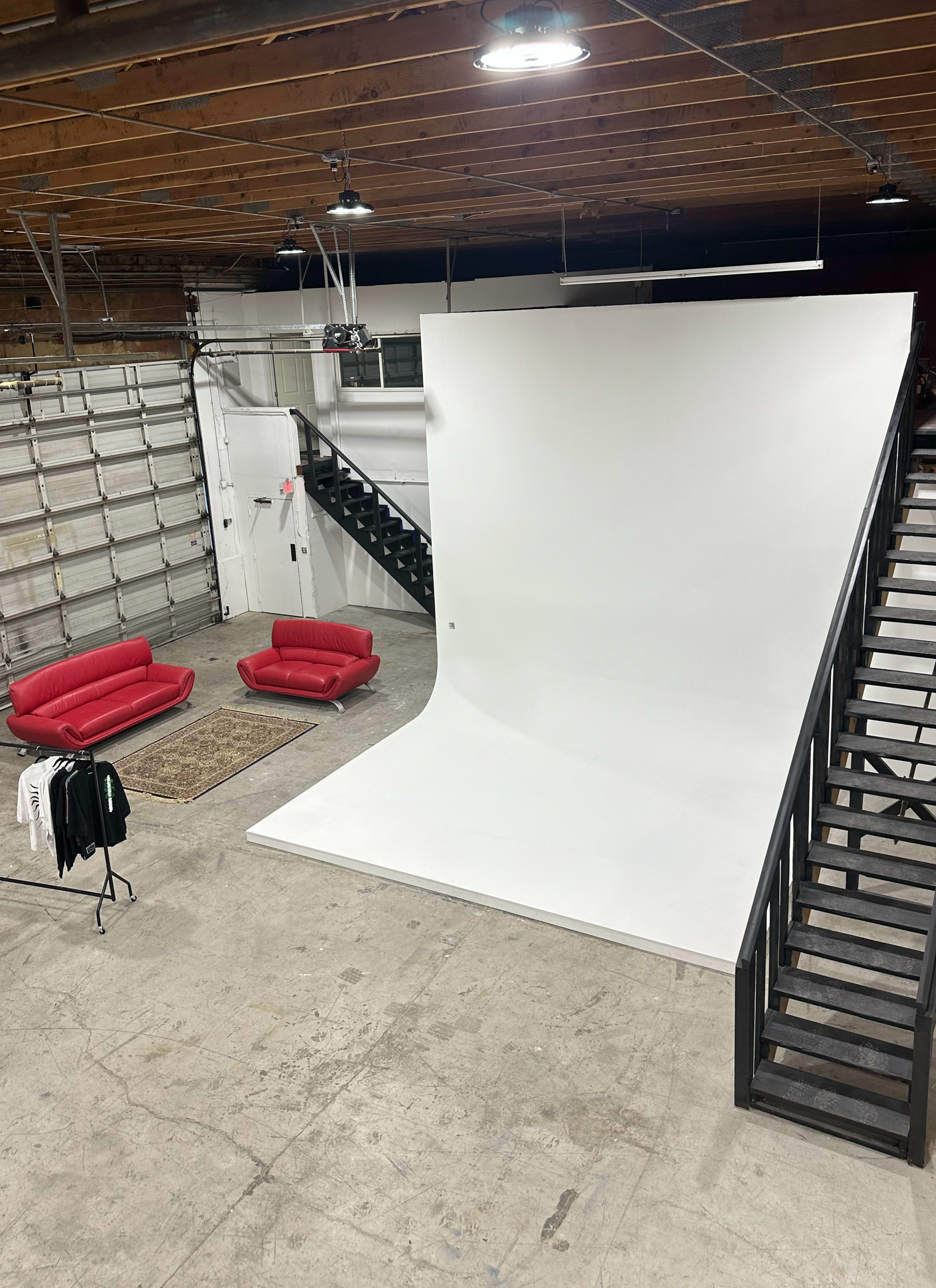 The image shows a spacious studio featuring a large white backdrop, two red sofas, a staircase, and a rug on a concrete floor.