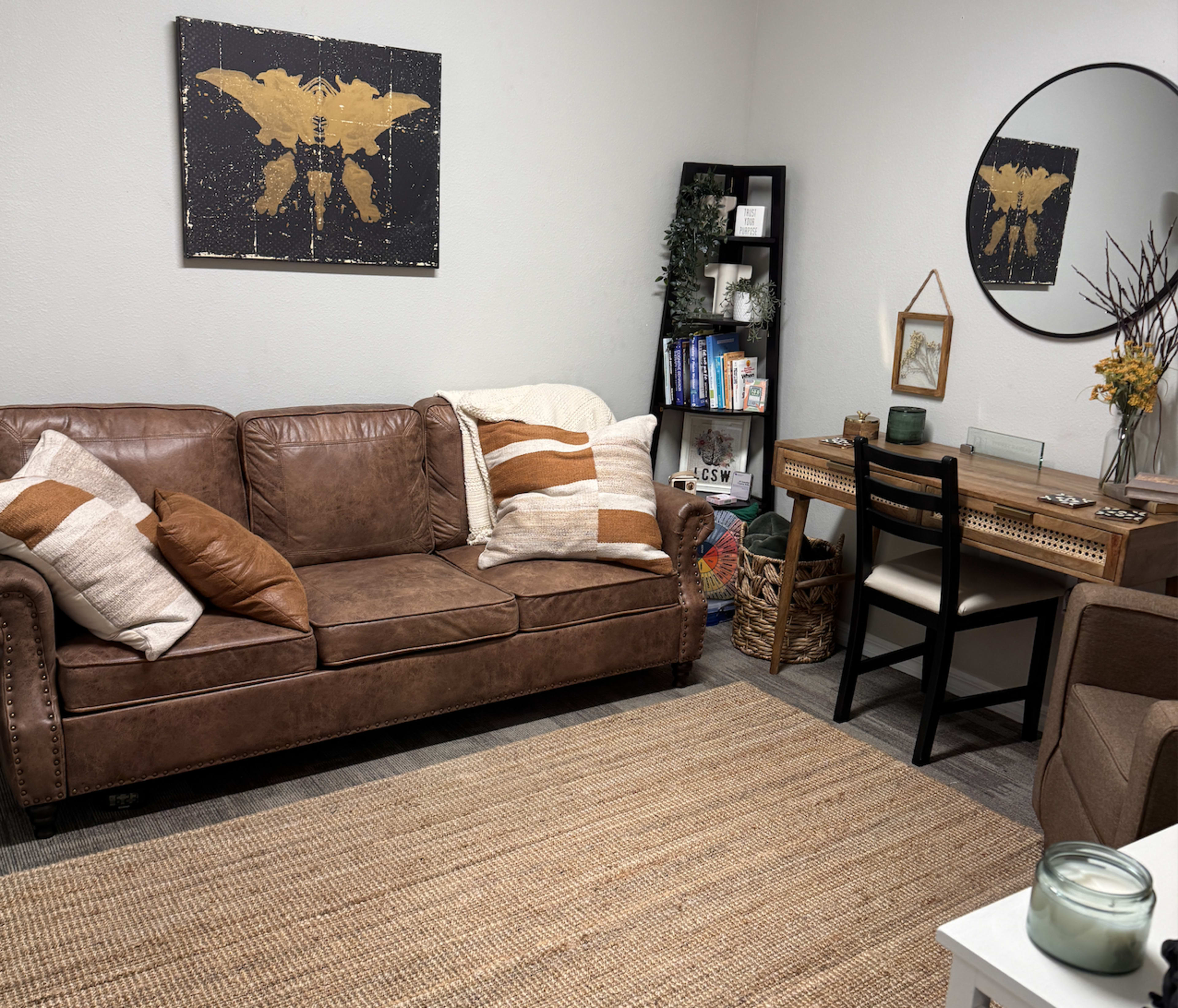 The image shows a cozy living room featuring a brown leather sofa with decorative pillows, a round mirror on the wall, a bookshelf filled with books, and a small wooden desk with a chair.