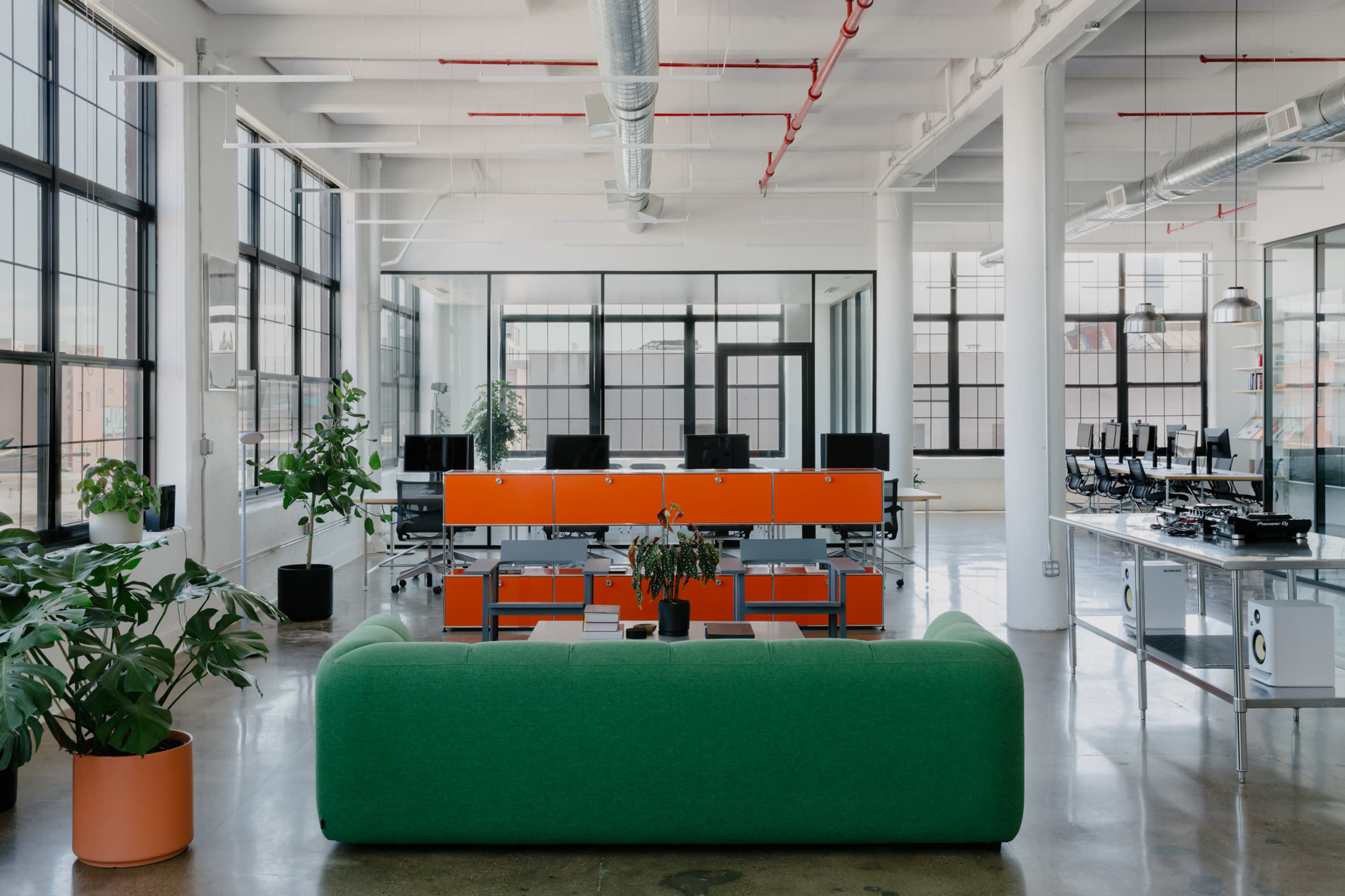 A bright, modern office space features green and orange furniture, large windows, and computer workstations arranged in a collaborative setup.