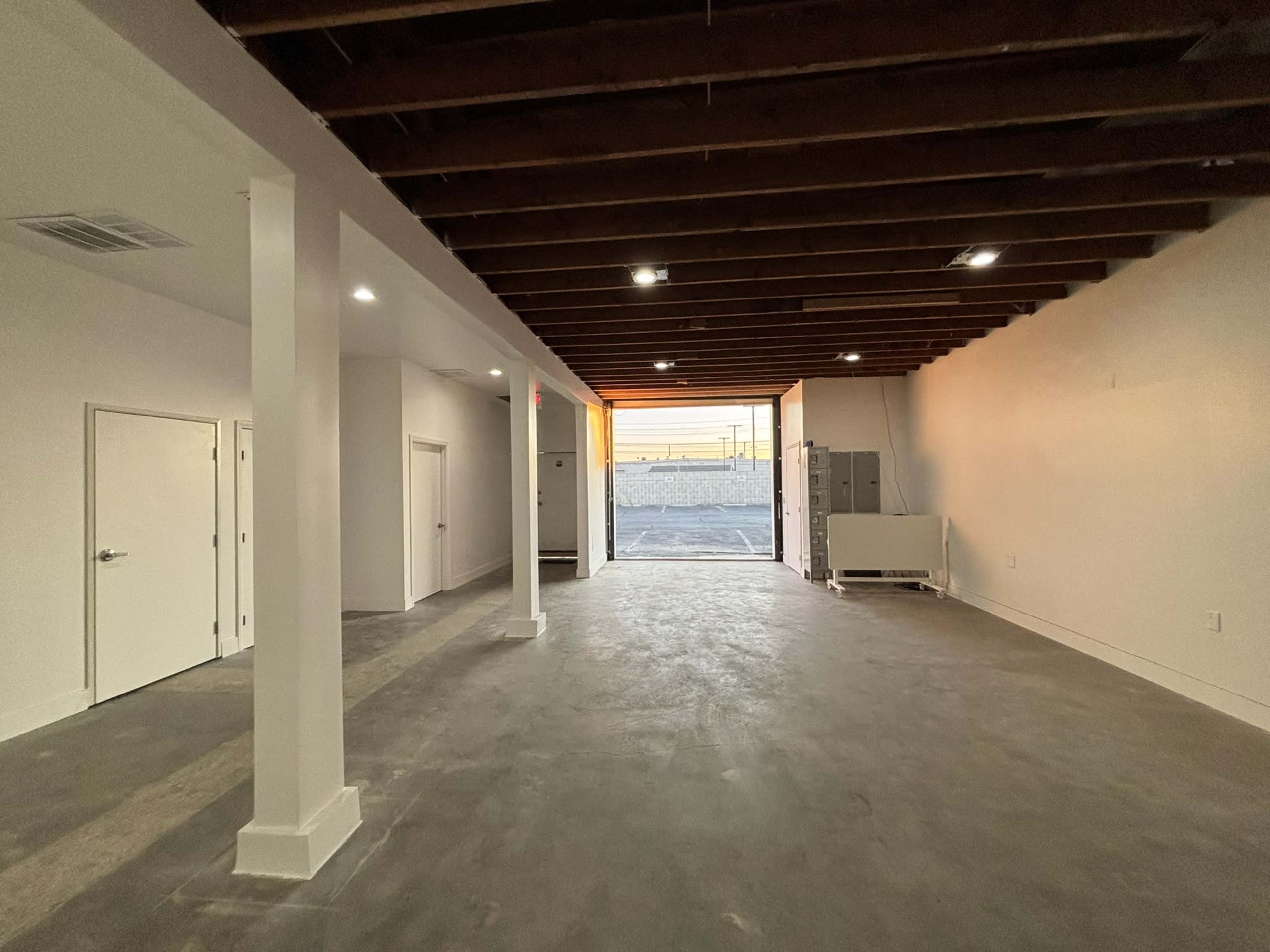 The image shows an empty room with exposed wooden beams on the ceiling, white walls, and a large window letting in natural light.