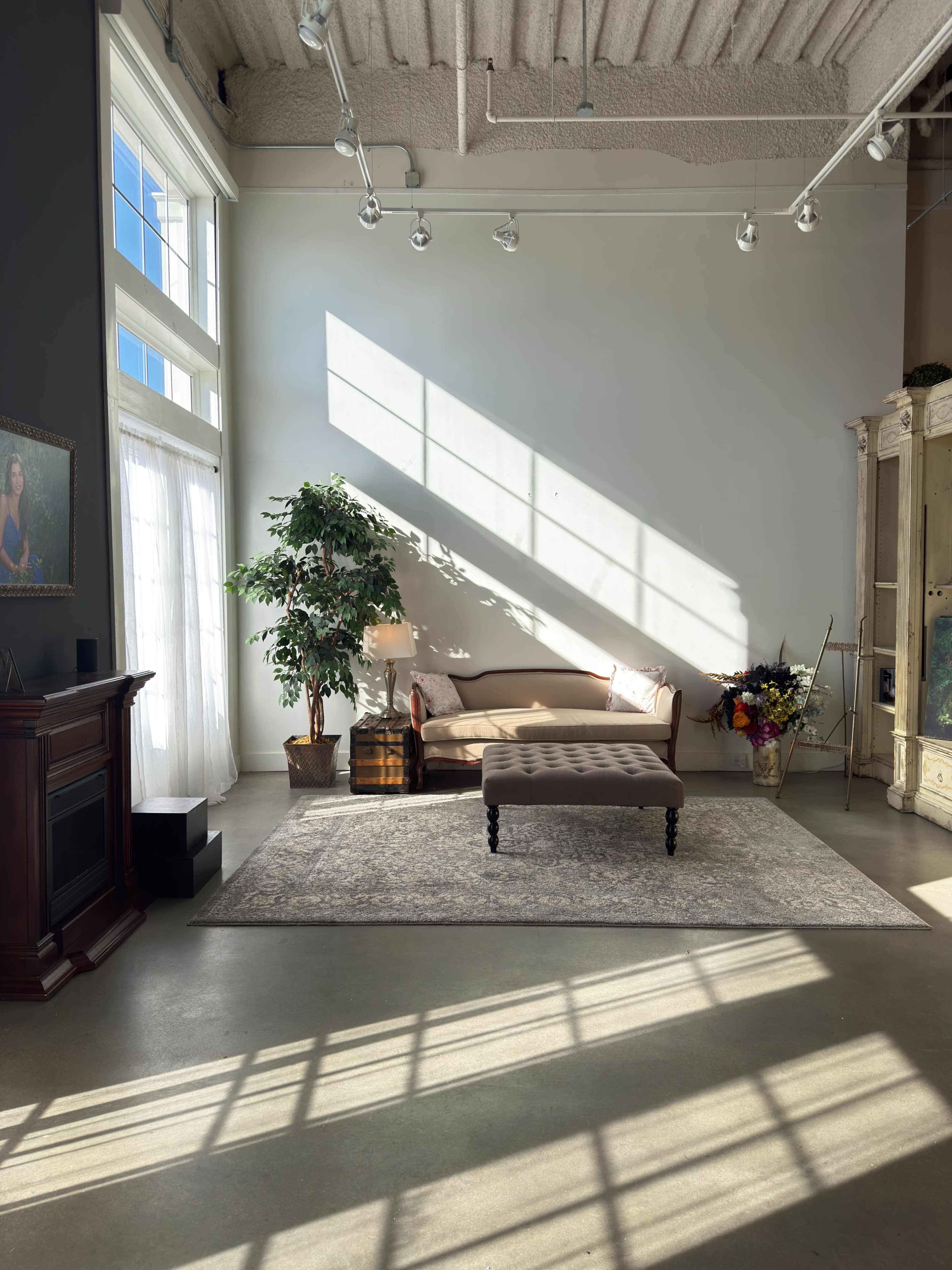 The image shows a well-lit room with a sofa, an ottoman, a potted plant, and a decorative cabinet, all highlighted by diagonal sunlight streaming through large windows.