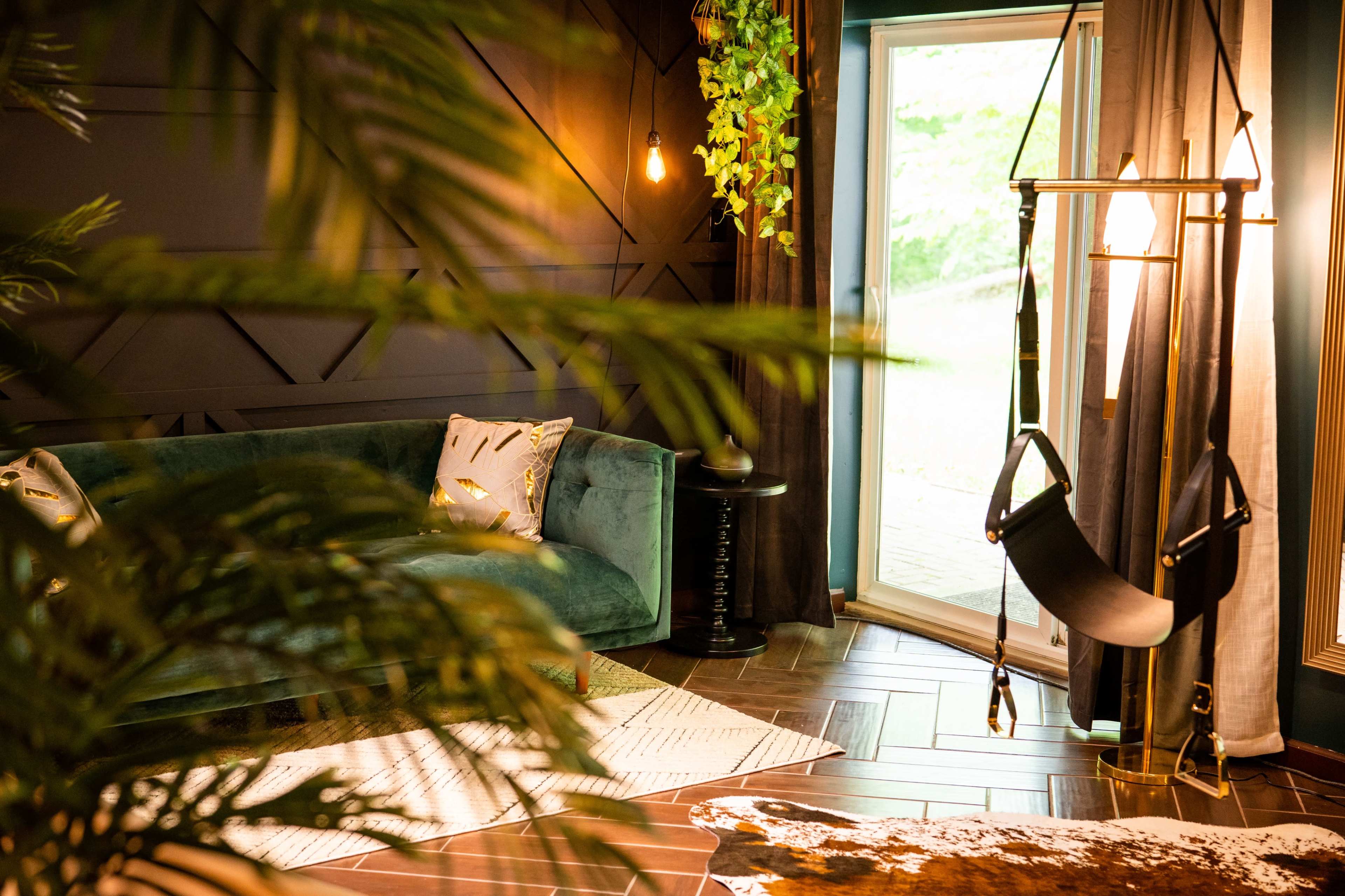 A stylish living room featuring a green velvet sofa, a suspended swing chair, and large windows with natural light streaming in.