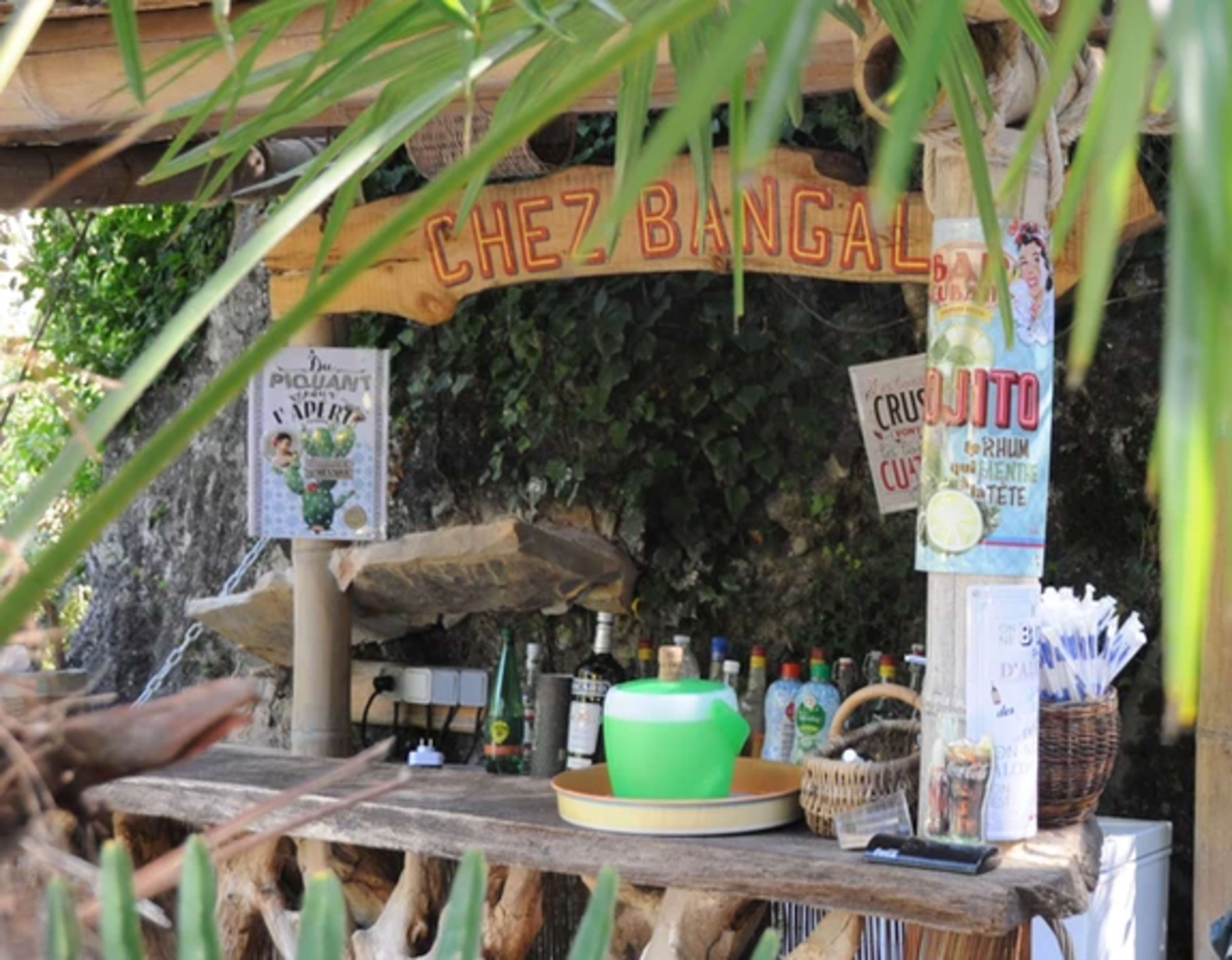 A rustic outdoor bar named "Chez Bangal" features a wooden counter with various bottles and a green bowl, surrounded by lush greenery.