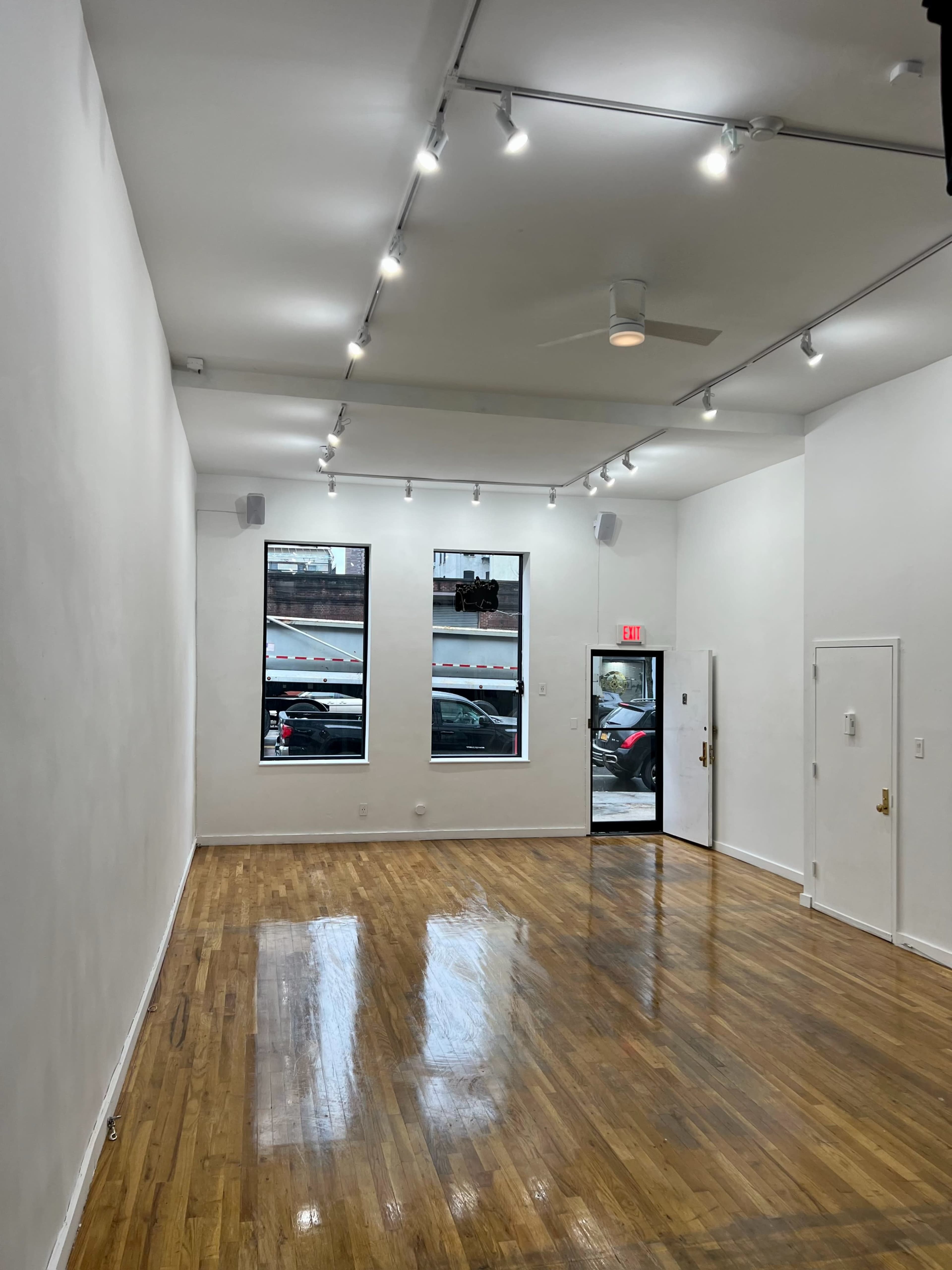 The interior of a spacious, well-lit room with white walls and glossy wooden floors features large windows and a door leading to the street.