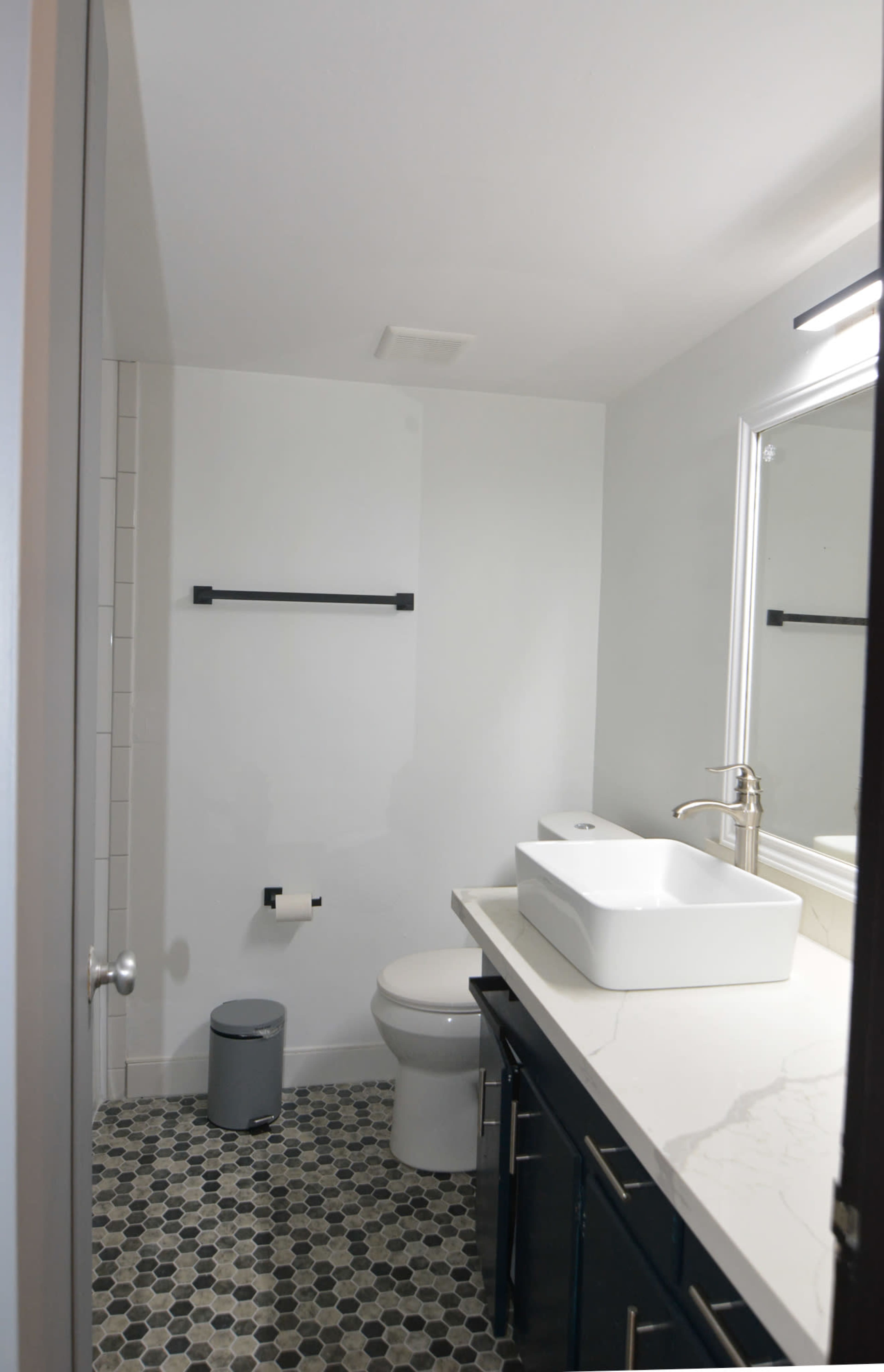 A modern bathroom with a white vessel sink on a countertop, a toilet, a gray trash can, and hexagonal tile flooring.