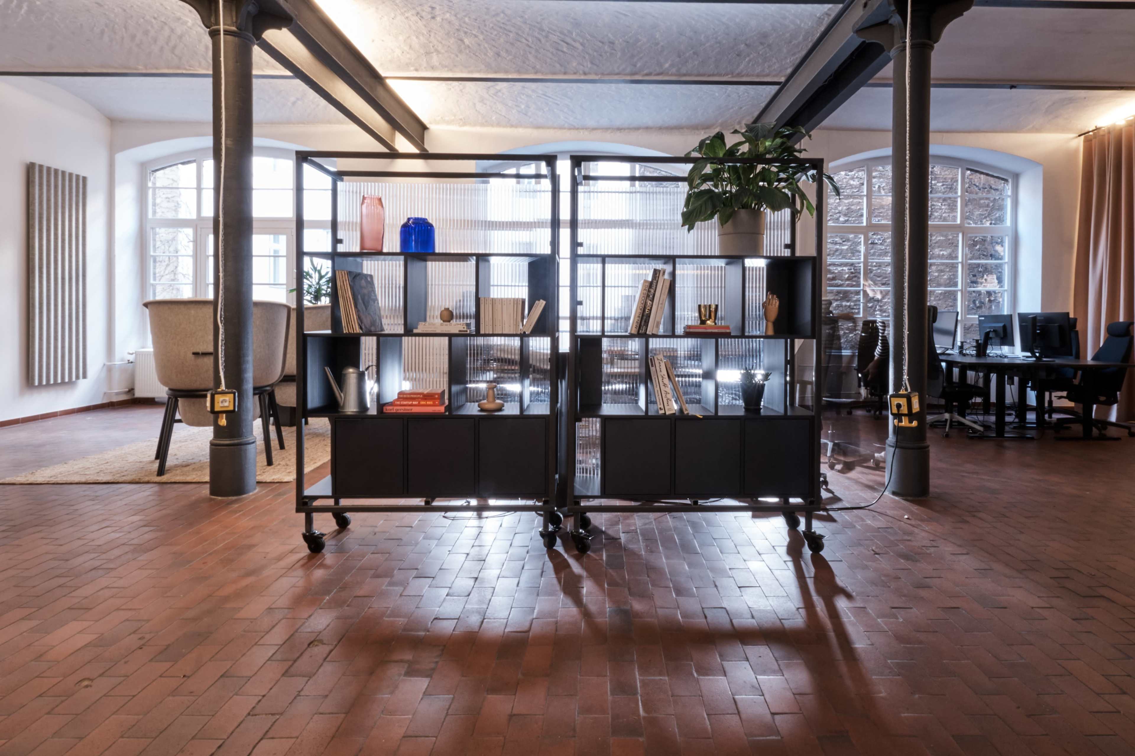 The image shows a modern office space with a metal bookshelf on wheels, filled with books and decorative items, separated from a work area featuring desks and chairs.