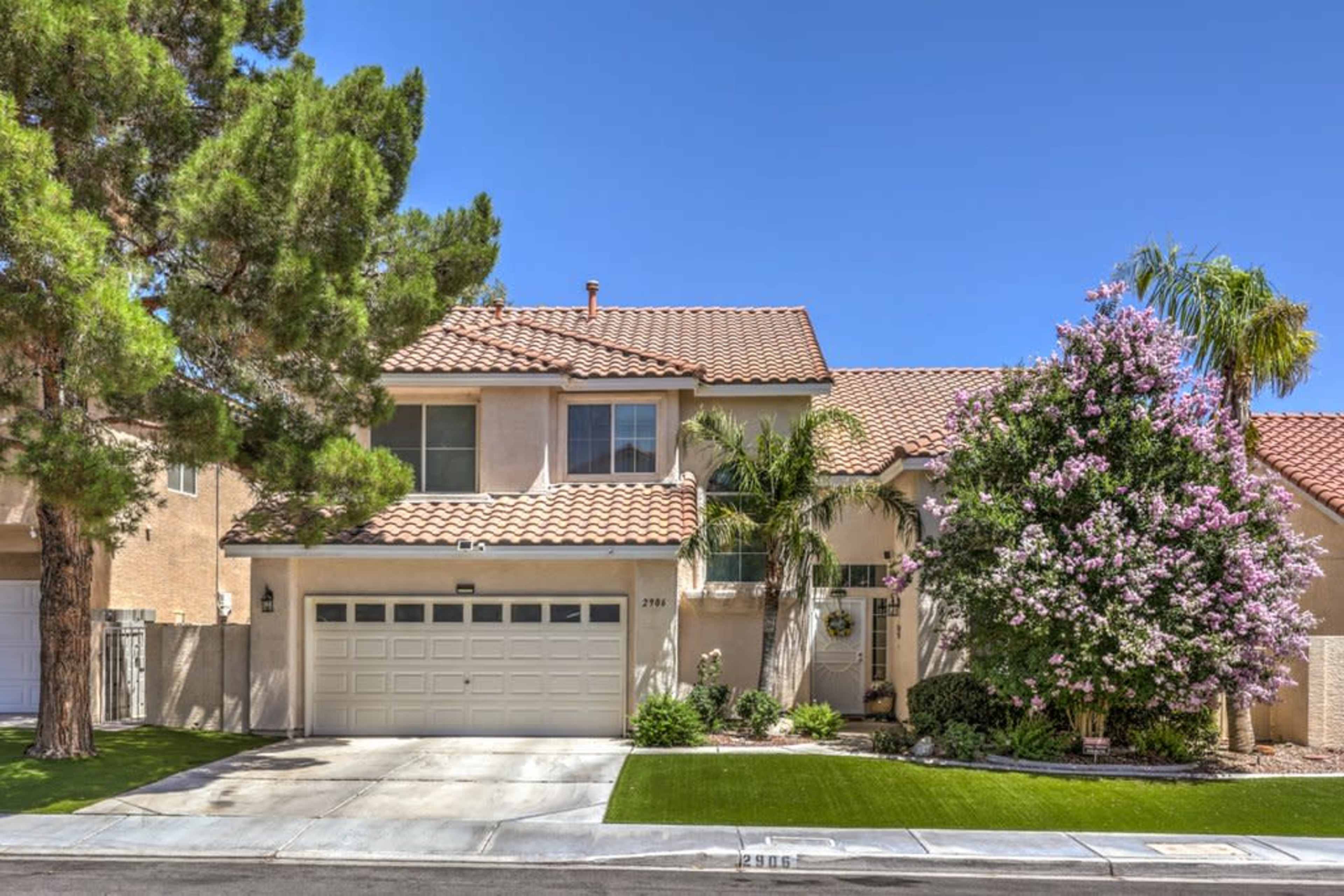 The image shows a two-story house with a tiled roof, surrounded by greenery and a flowering bush in the front yard.