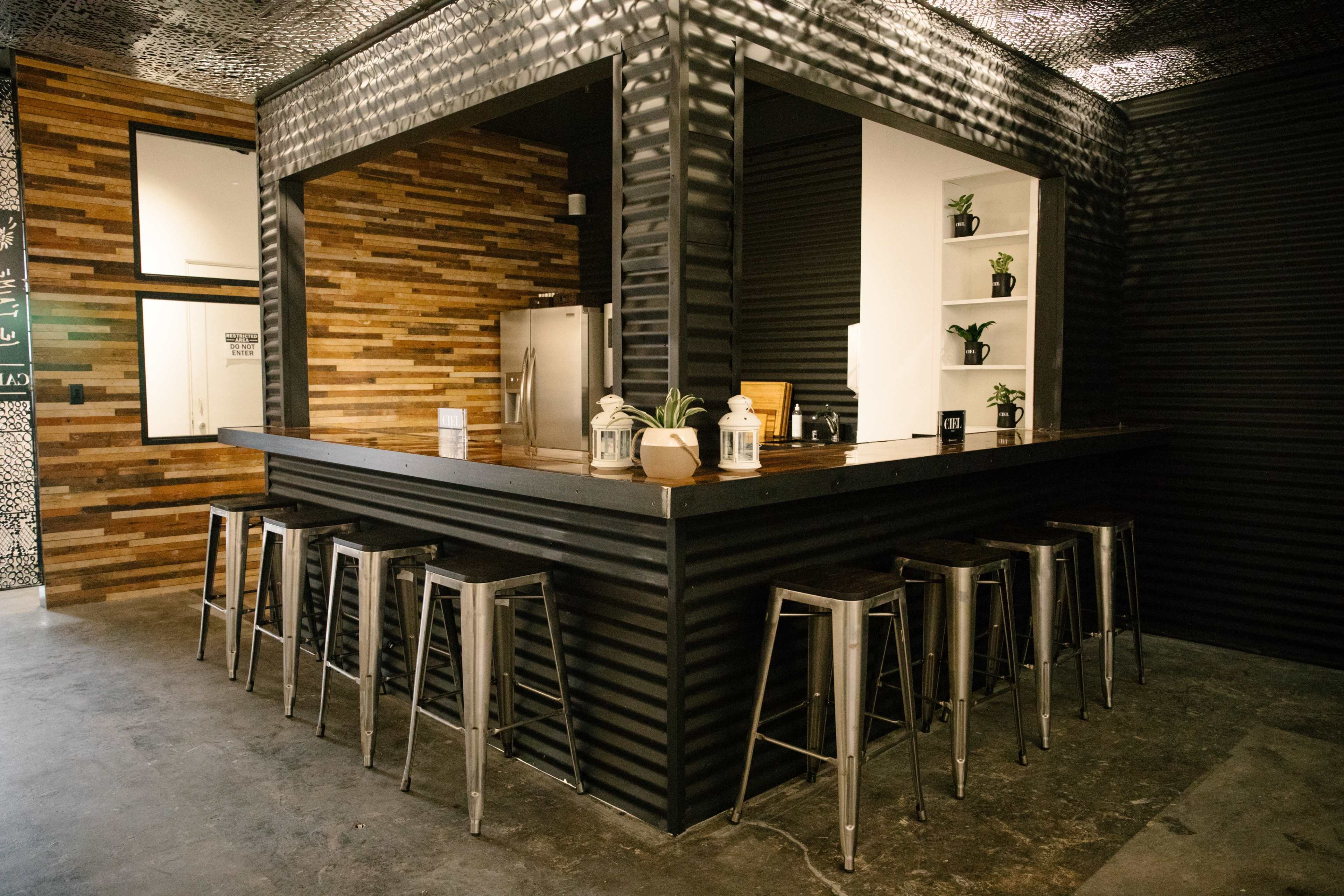 A modern kitchen with a black corrugated metal bar area, wooden walls, stainless steel stools, and potted plants.