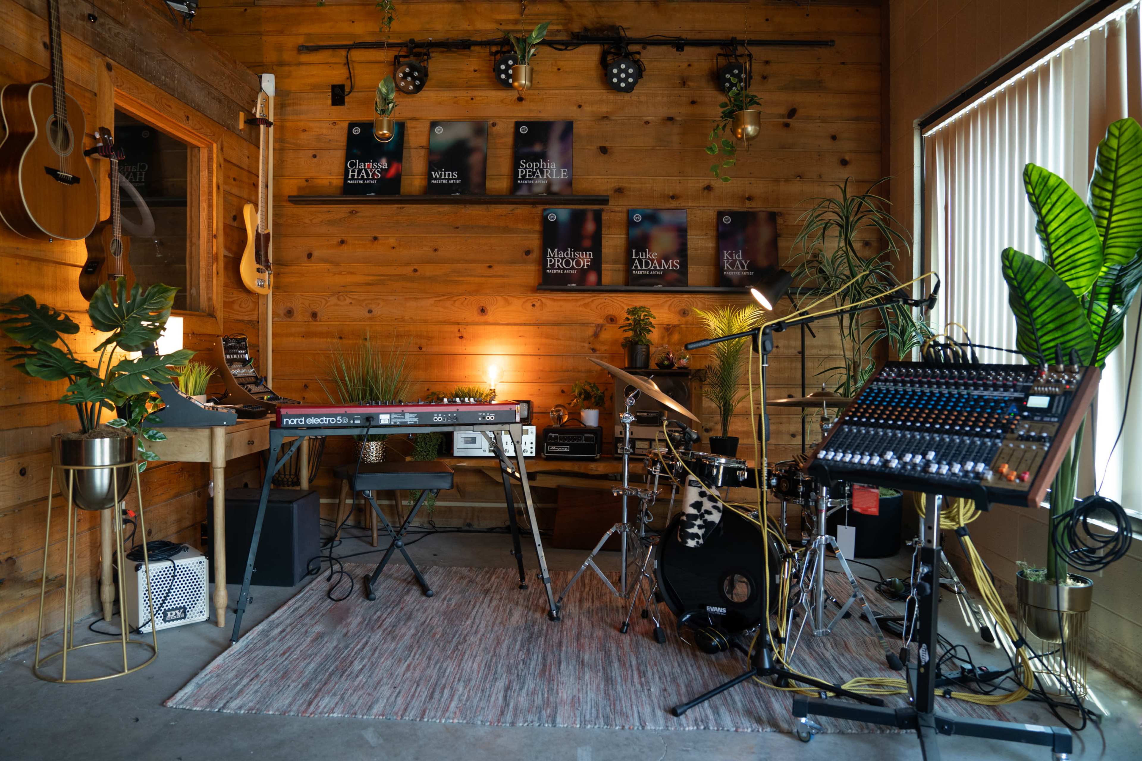 A music studio featuring various instruments, including a keyboard, drum set, microphones, and decorative plants, with wooden walls and framed posters.