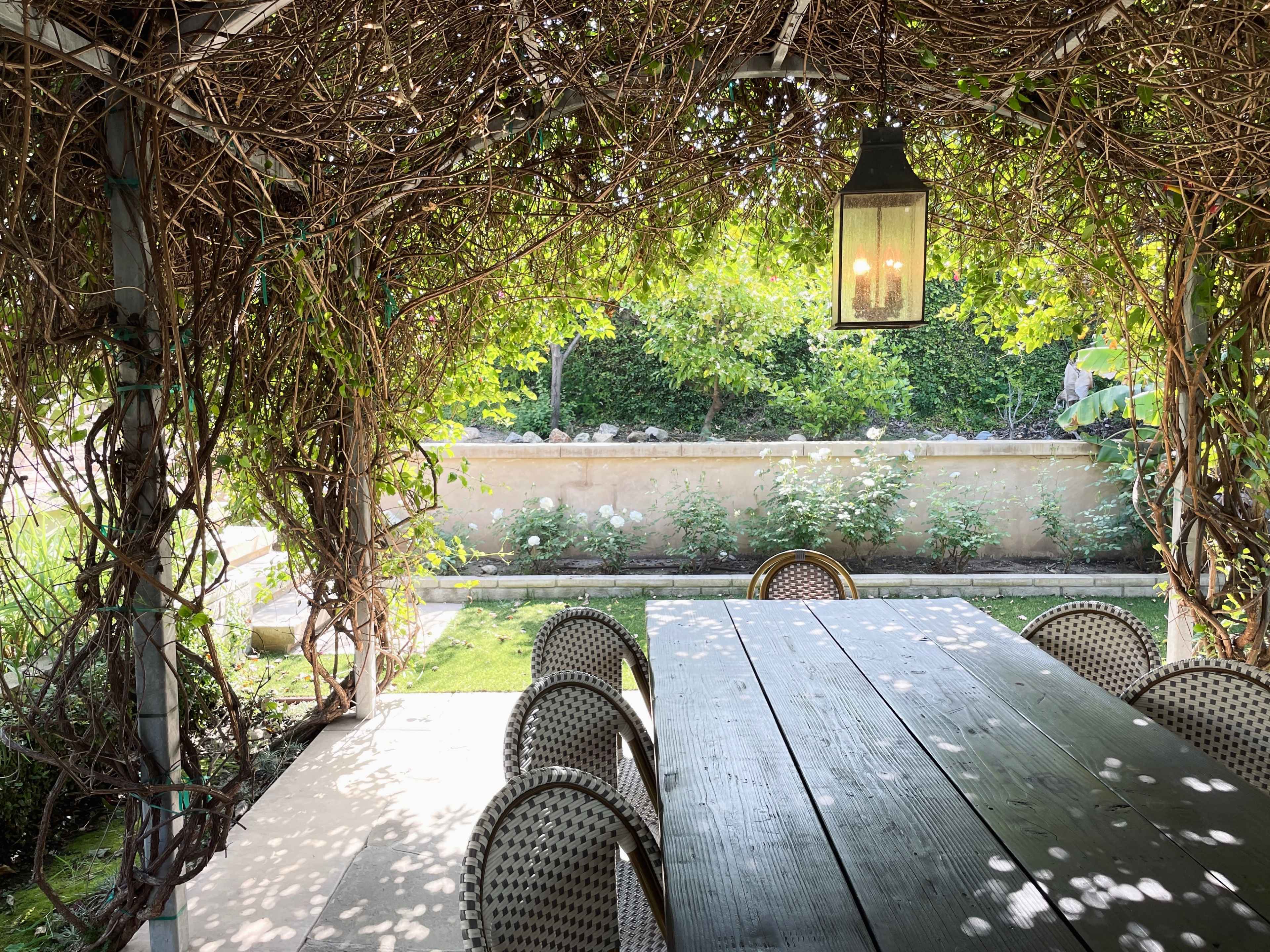 A shaded outdoor dining area features a wooden table surrounded by chairs, with a lantern overhead and greenery visible in the background.
