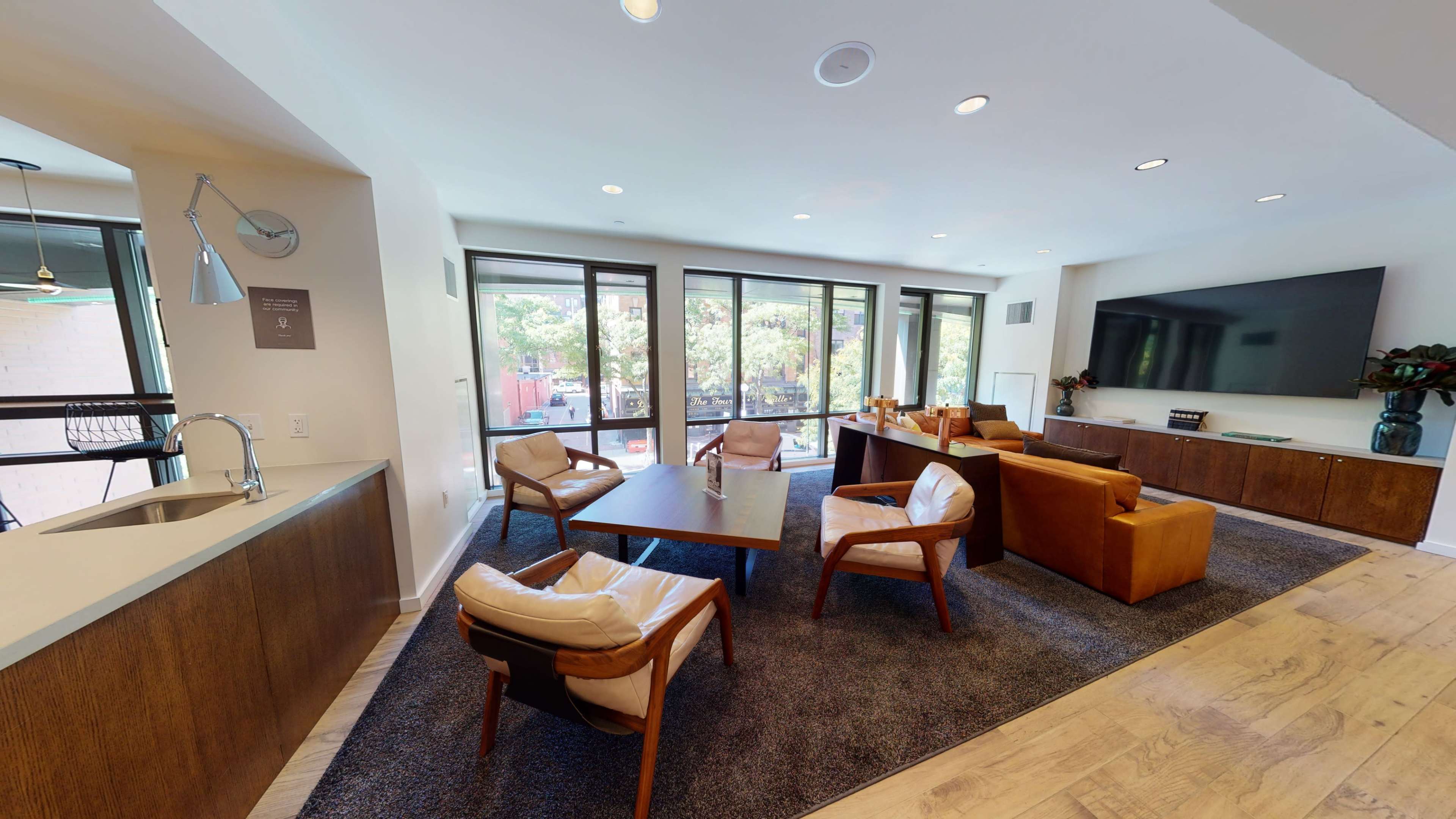 A modern living area featuring a comfortable seating arrangement with chairs and a table, large windows providing natural light, and a wall-mounted television.